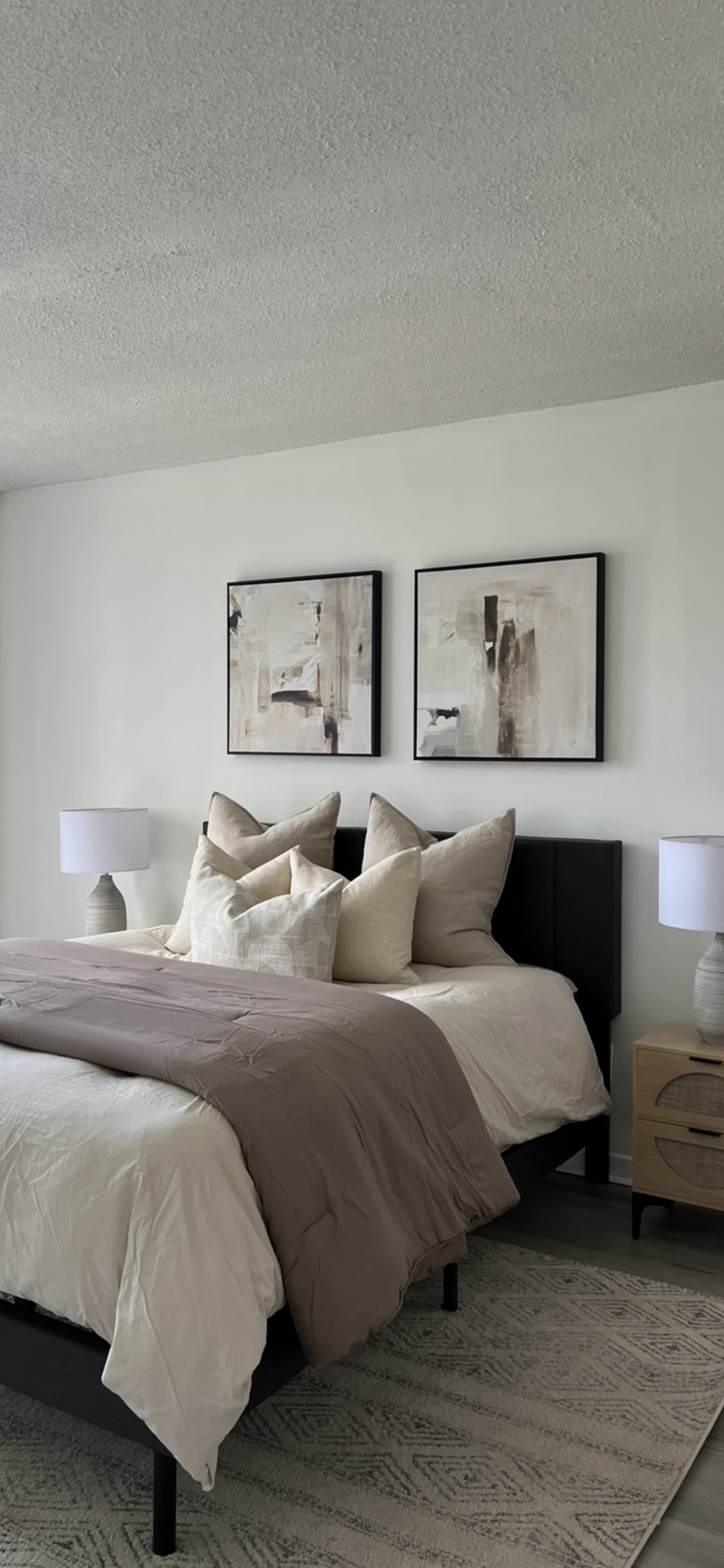 A neatly made bed with beige and cream pillows, a taupe comforter, and two white lamps on nightstands in a modern bedroom with abstract artwork on the wall.