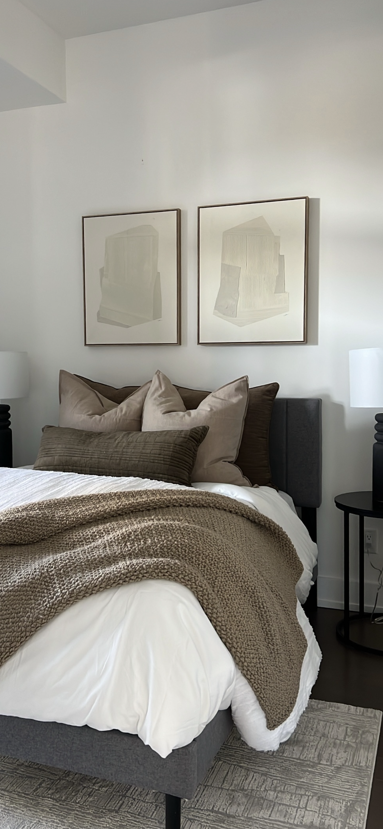 A neatly made bed with multiple pillows, a beige blanket, and a white duvet in a contemporary bedroom, with two framed abstract art pieces above the headboard and two bedside lamps.