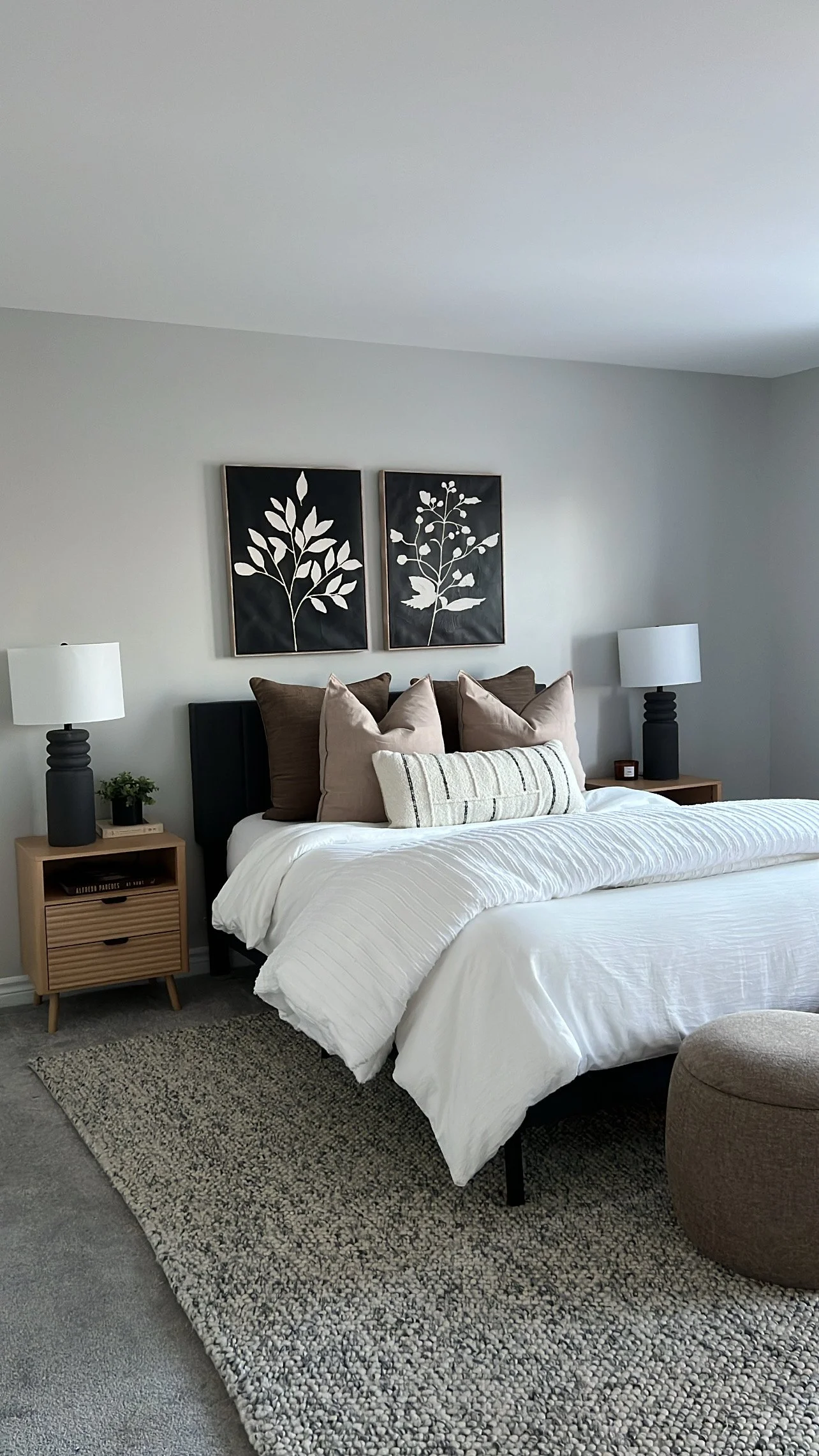 A neatly made bed with white bedding and multiple pillows in front of black headboard, flanked by two wooden nightstands with black lamps and small plants, with two black and white floral art pieces hanging above the bed.