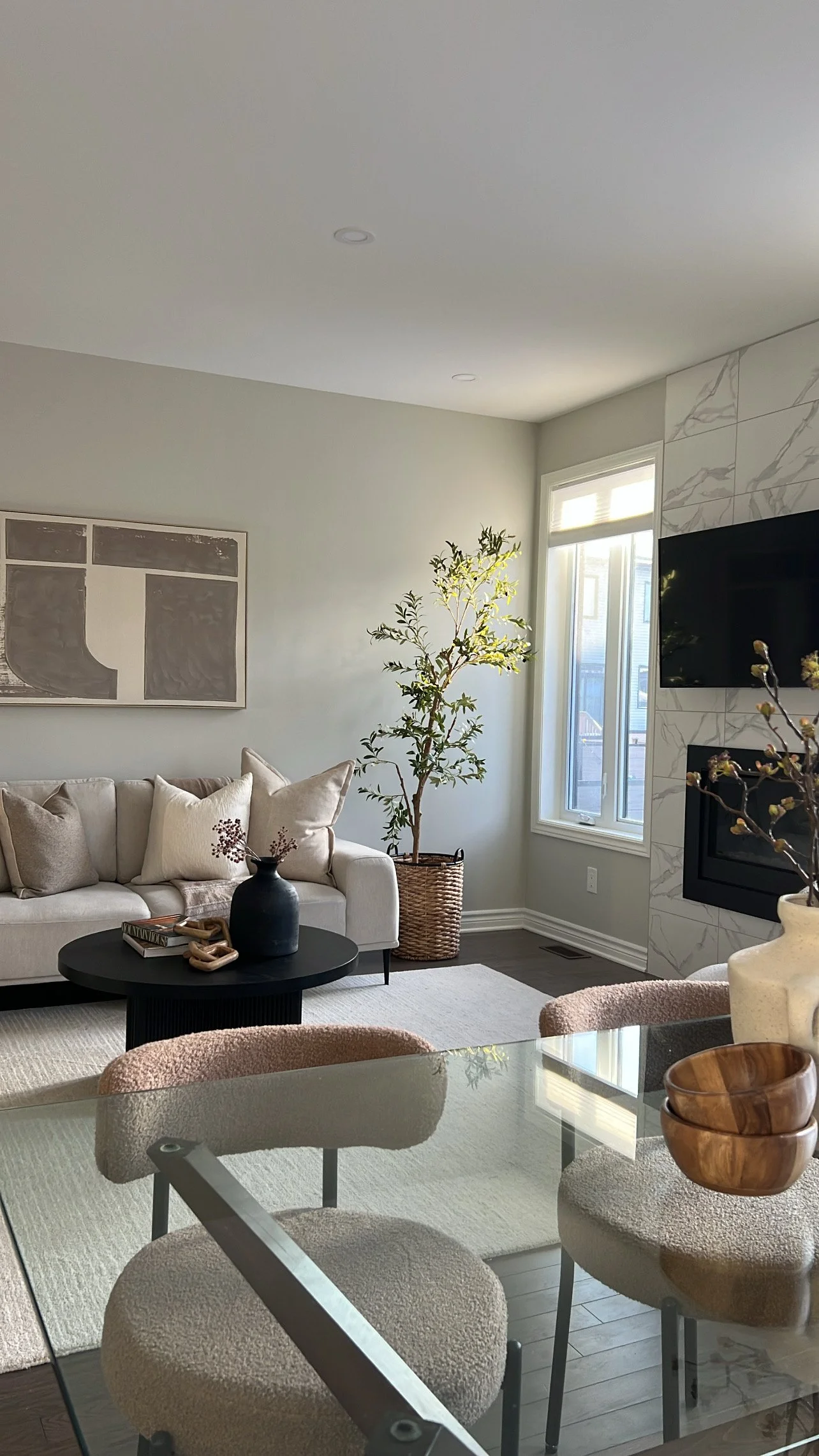 Living room with beige sofa, throw pillows, black coffee table with decorative items, large window, potted plant, wall art, and fireplace.