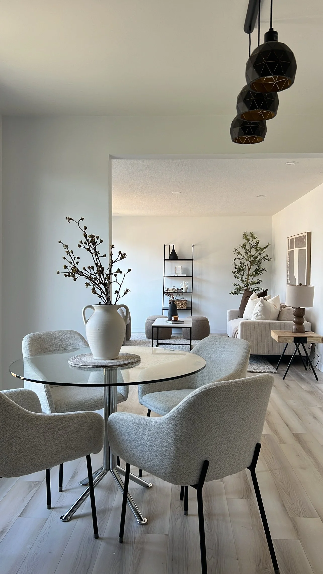 A modern living room and dining area with light-colored furniture, a round glass table with four beige upholstered chairs, a large white ceramic vase with branches on the table, a white sofa with pillows, a wooden side table with a lamp, a black meta