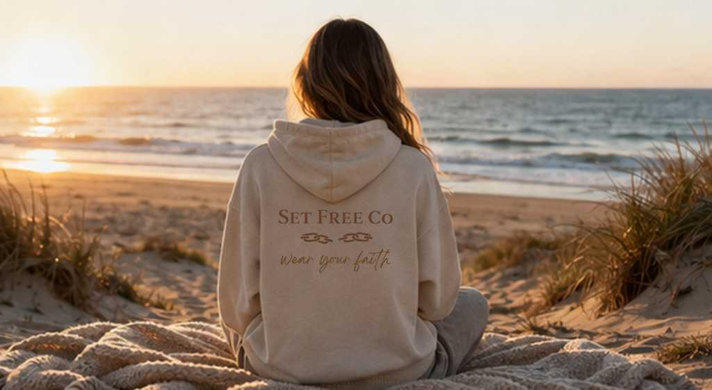 Woman sitting on beach at sunset, facing the ocean, wearing a beige hoodie with the words "Set Free Co" and "wear your faith" printed on the back.