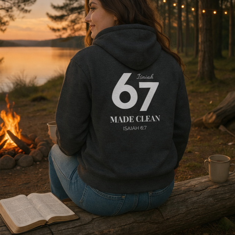 A woman sitting on a log near a campfire and a lake at sunset, wearing a dark hoodie with the text '67 Isaiah Made Clean' on the back. There are string lights hanging in the background and an open Bible beside her.