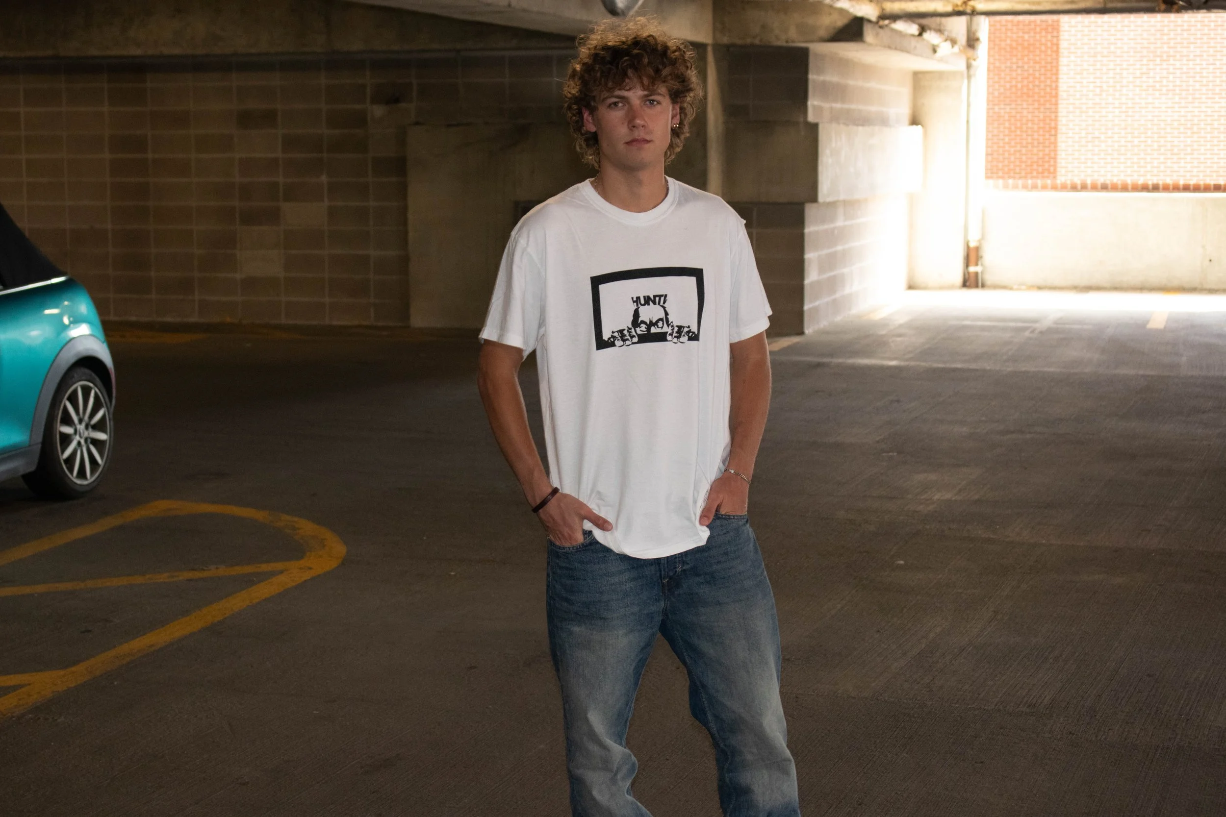 A young man with curly hair wearing a white T-shirt with a black graphic and blue jeans, standing in an underground parking garage.