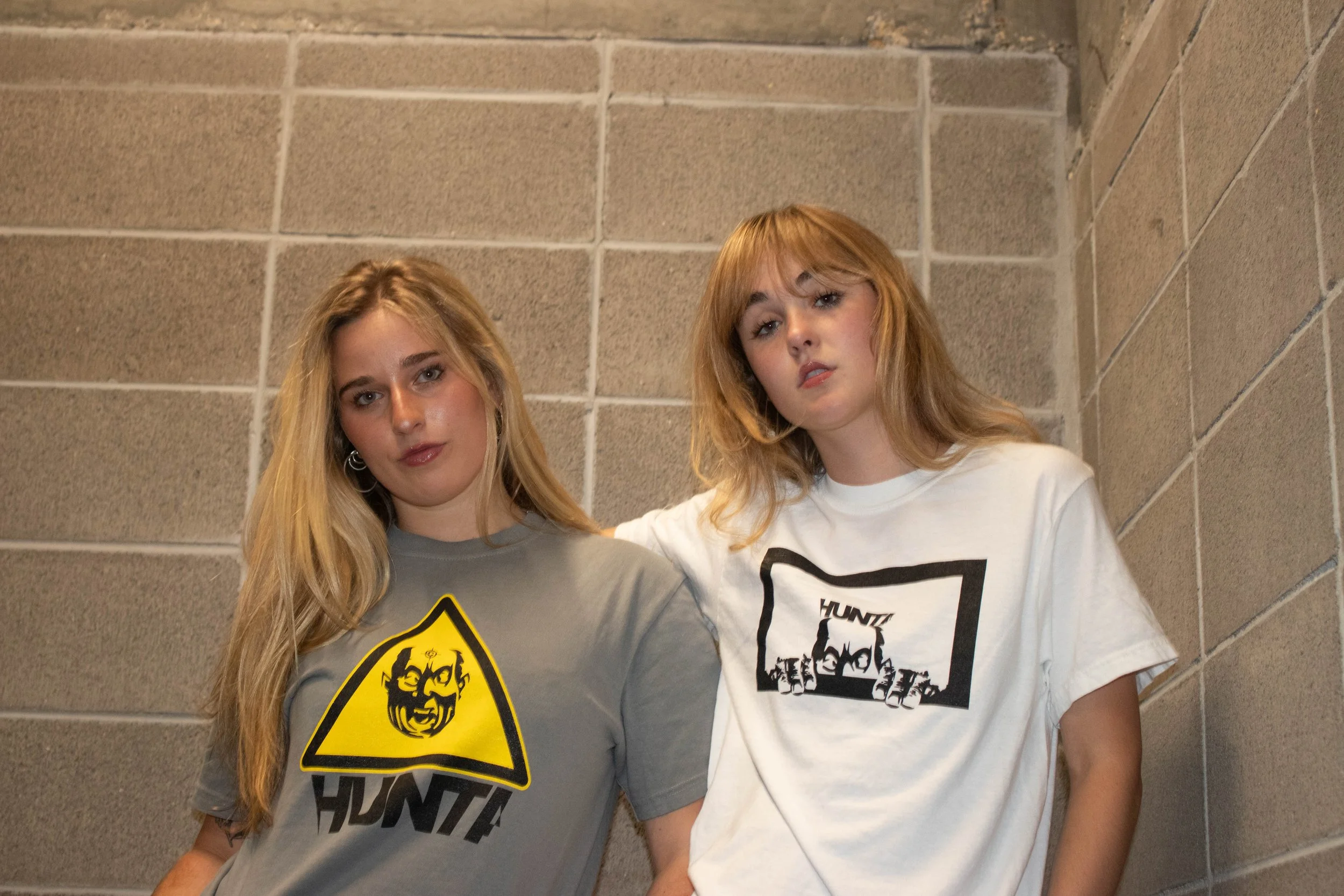 Two young women with blonde hair standing together against a beige stone wall, wearing T-shirts with graphic designs.