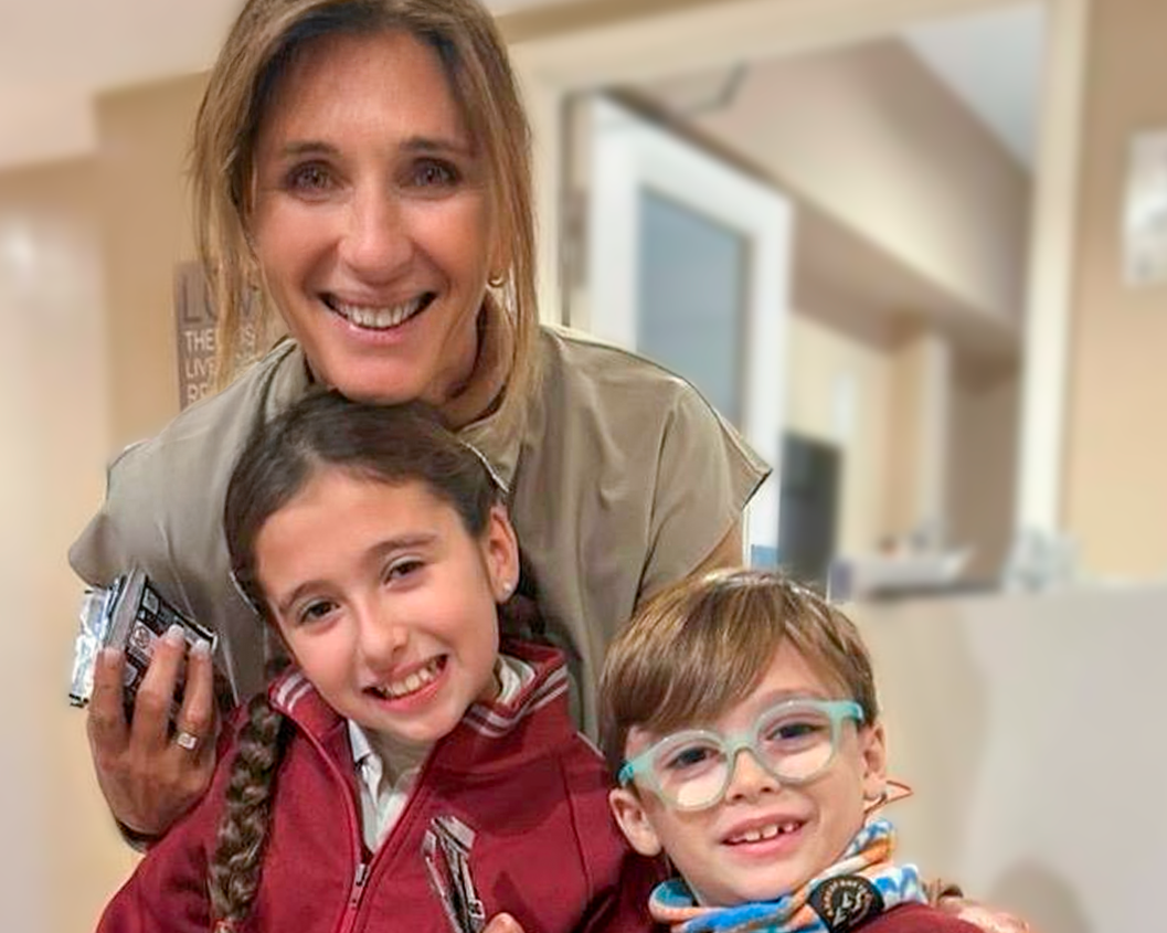 Una mujer y dos niños pequeños sonriendo en un interior, con la mujer en el centro y los niños a su lado. Todos parecen felices y están posando para la foto.