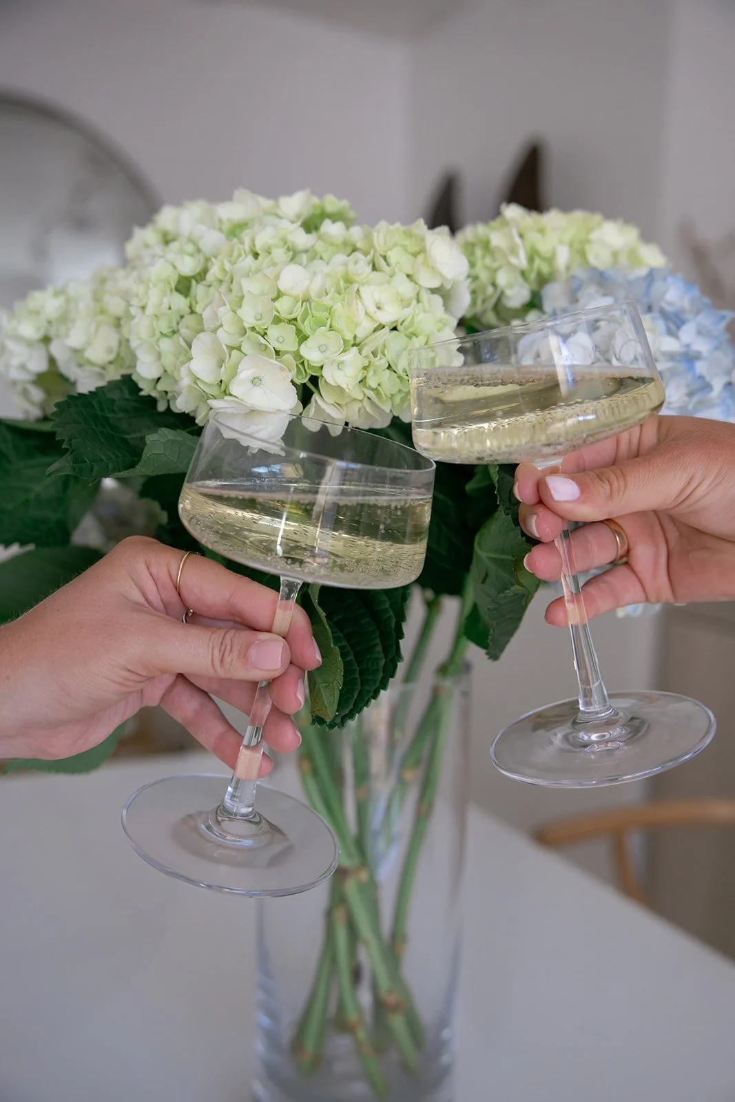 Two people holding champagne glasses clinking in front of a bouquet of white and pale green flowers.