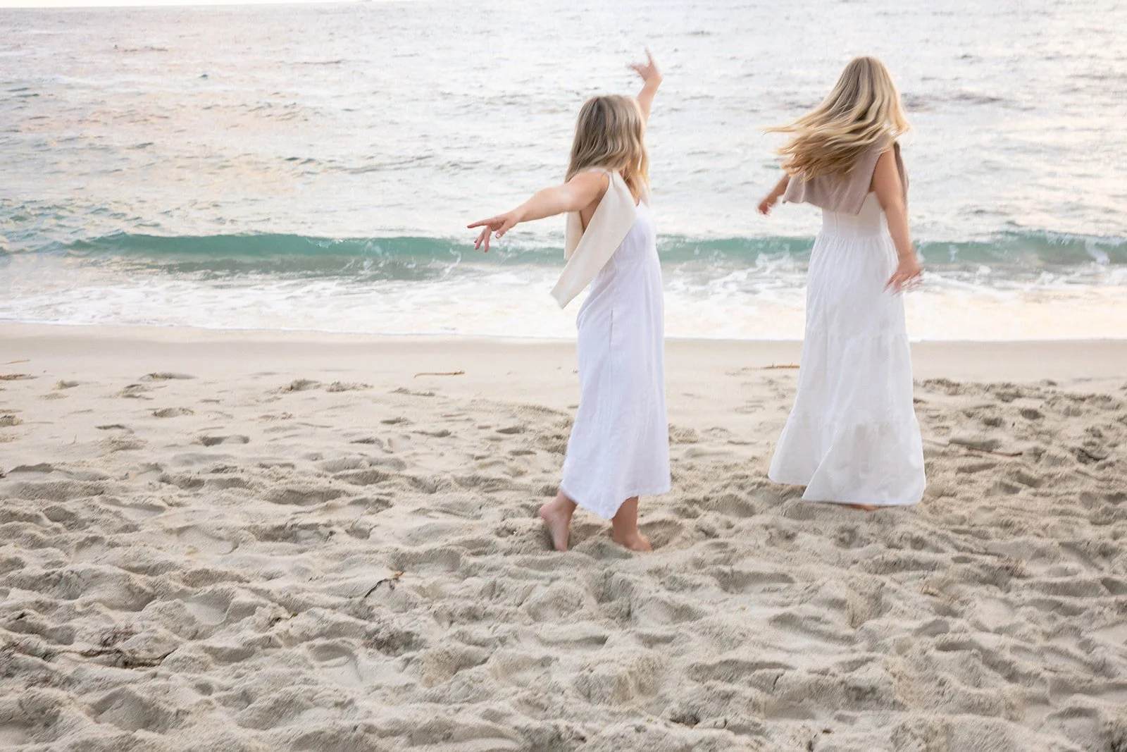 Two women in white dresses walking on the sandy beach near the ocean, with one woman raising her arm and the other looking down, during sunset.