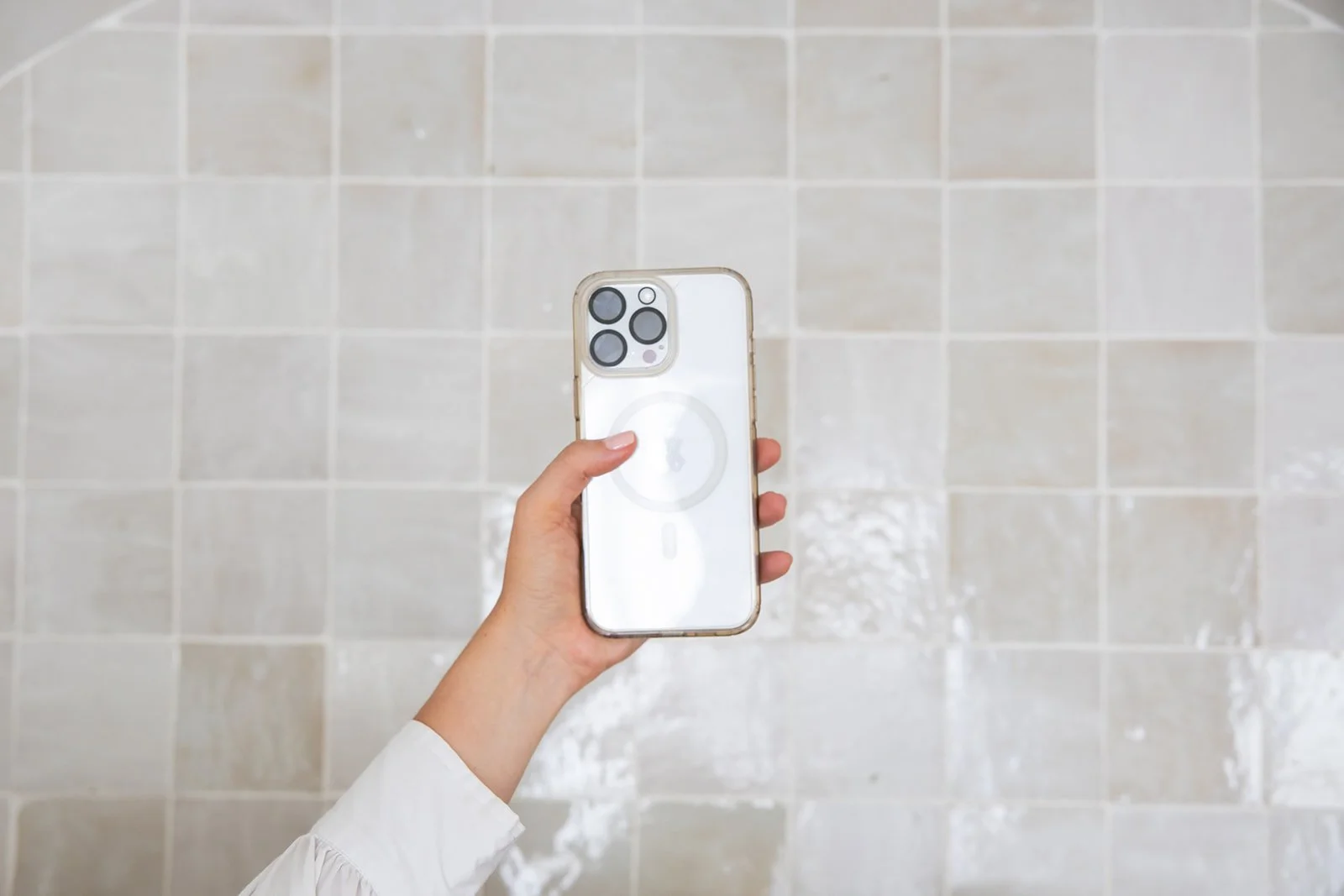Person holding an iPhone with a circle MagSafe charger attached, against a tiled bathroom wall.