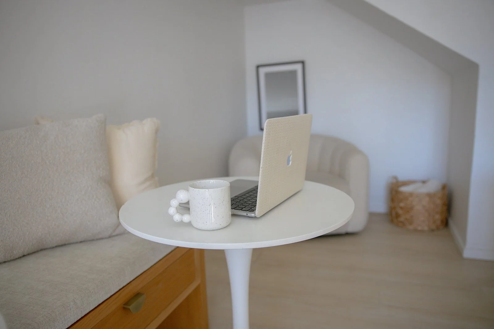 A white round table with a speckled white mug and a beige laptop on top. In the background, there is a beige sofa with textured cushions, a framed mirror on the wall, and a woven basket in the corner. The room has light-colored walls and wooden flooring.