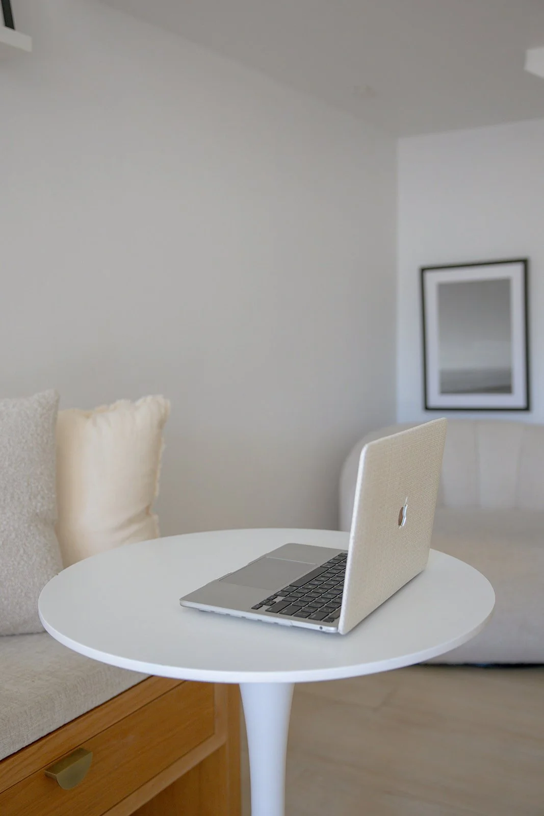 A white round table with a beige laptop on it, beside a beige sofa with fluffy pillows, in a minimalist living room with a framed picture on the wall.