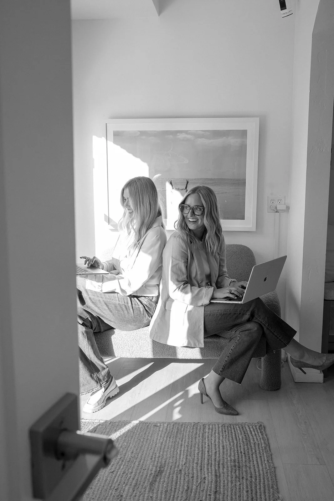 Two women sitting on a sofa, working on laptops, smiling, in a bright room.