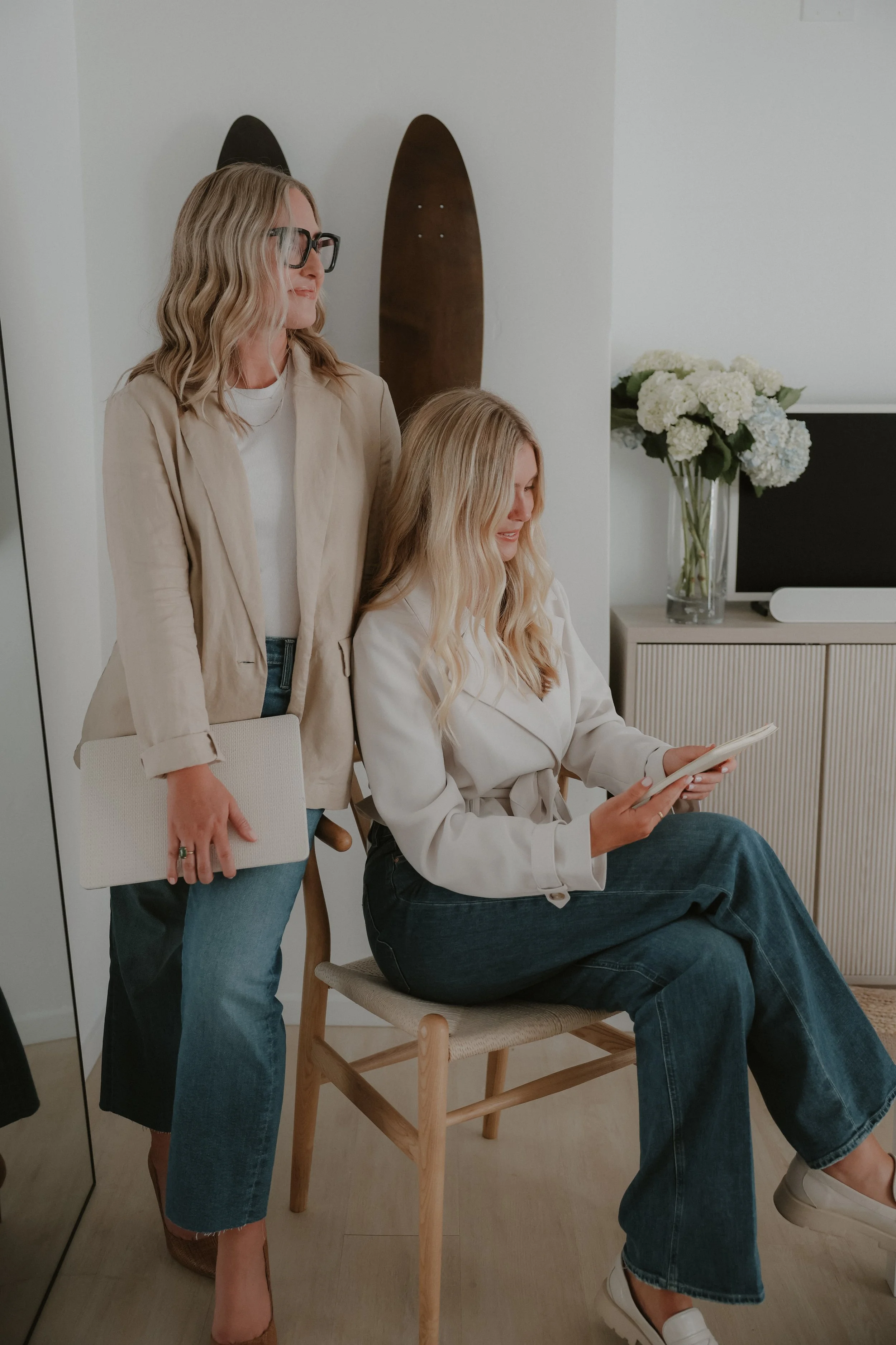 Two women with blonde hair, one sitting on a chair looking at a tablet, the other standing beside her holding a beige folder, in a modern room with a vase of white flowers and a black television.