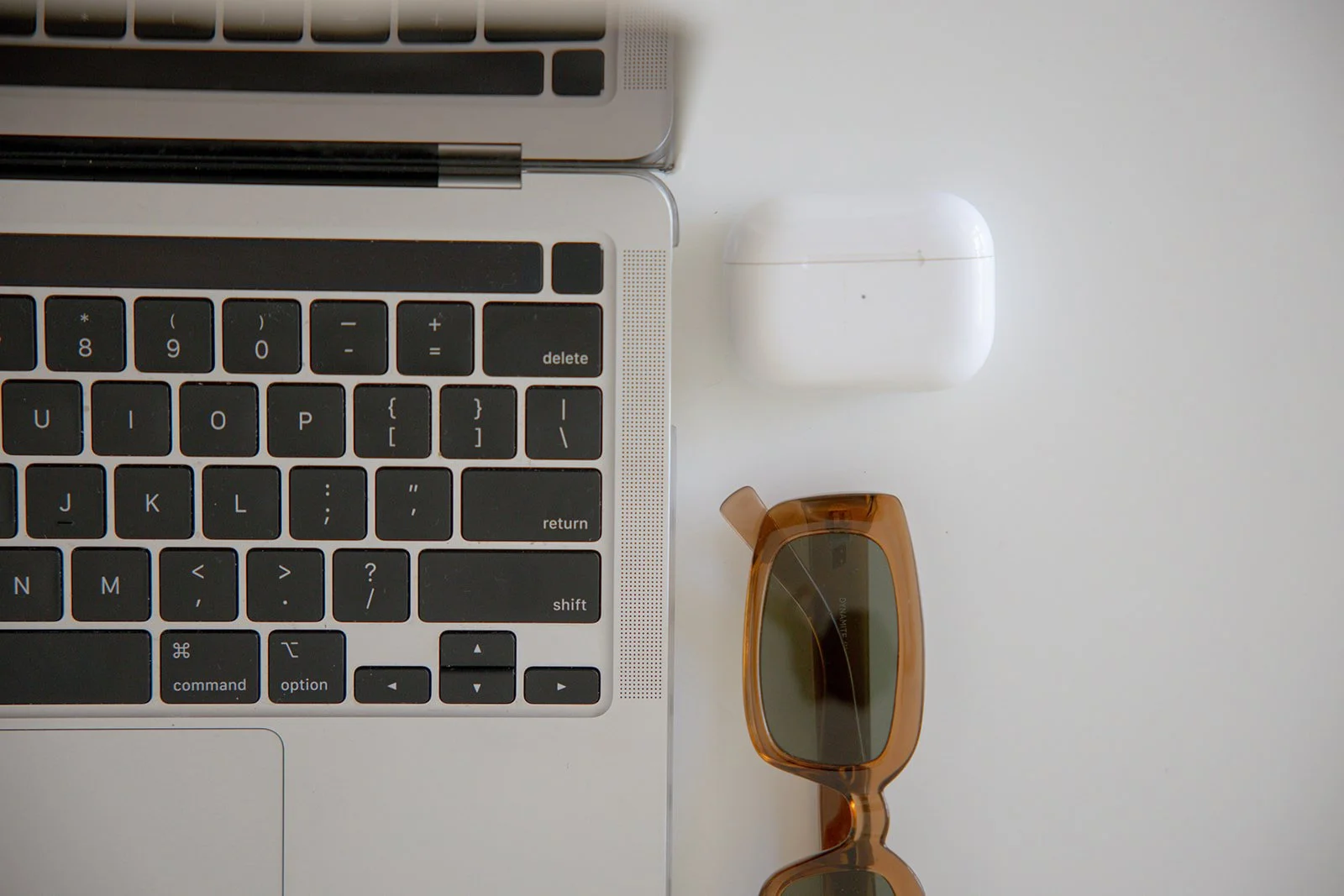 top left corner of a silver laptop with black keys, next to a white AirPods case, and below the case, a pair of brown sunglasses with dark lenses