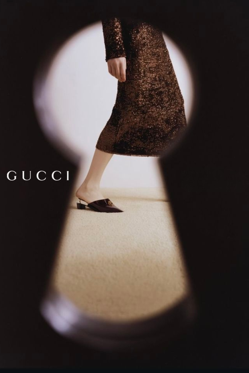 Part of a woman wearing a sparkly, sequined, brown dress and black Gucci mule shoes, walking.