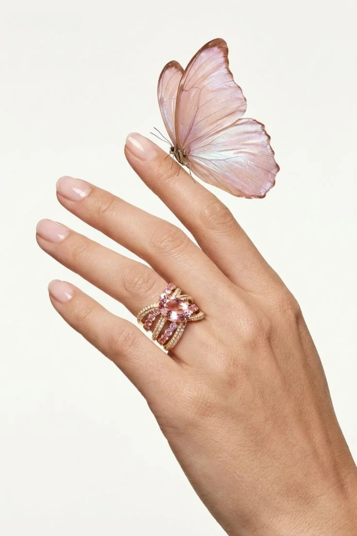 A close-up of a hand wearing multiple gold rings with pink gemstones, with a pink butterfly resting on the index finger against a plain white background.