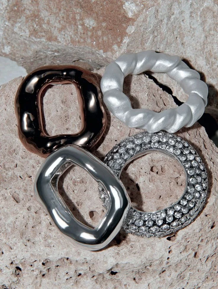 Four statement rings with different designs and finishes on a textured stone surface.