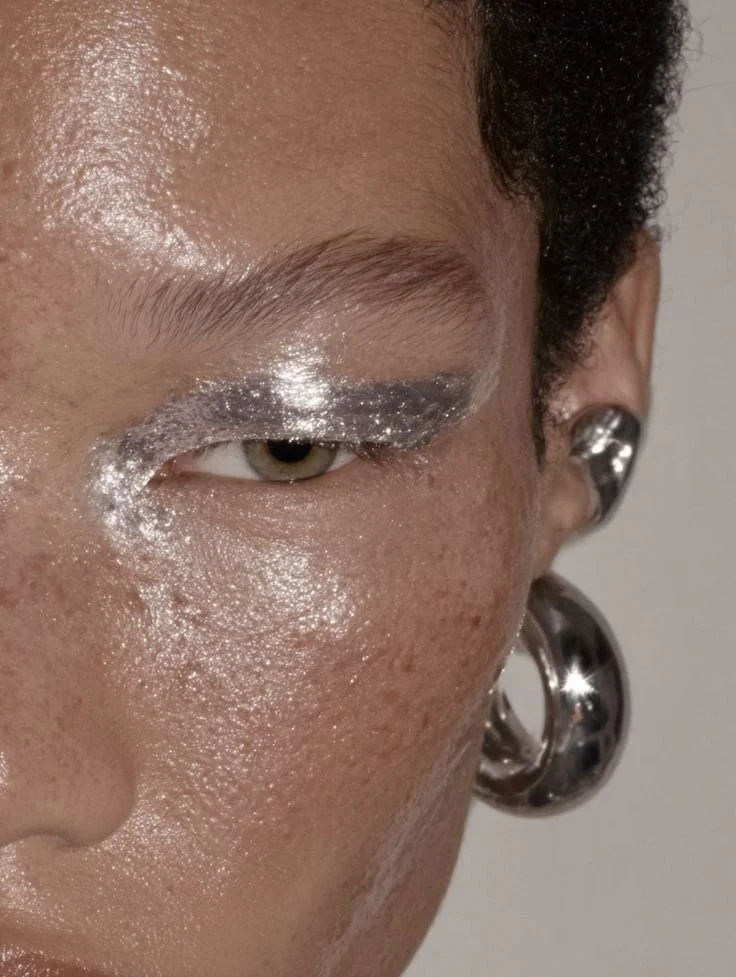 Close-up of a person's face with shiny, metallic silver makeup around the eyes, eyebrow, and cheekbone, and large, metallic earrings.