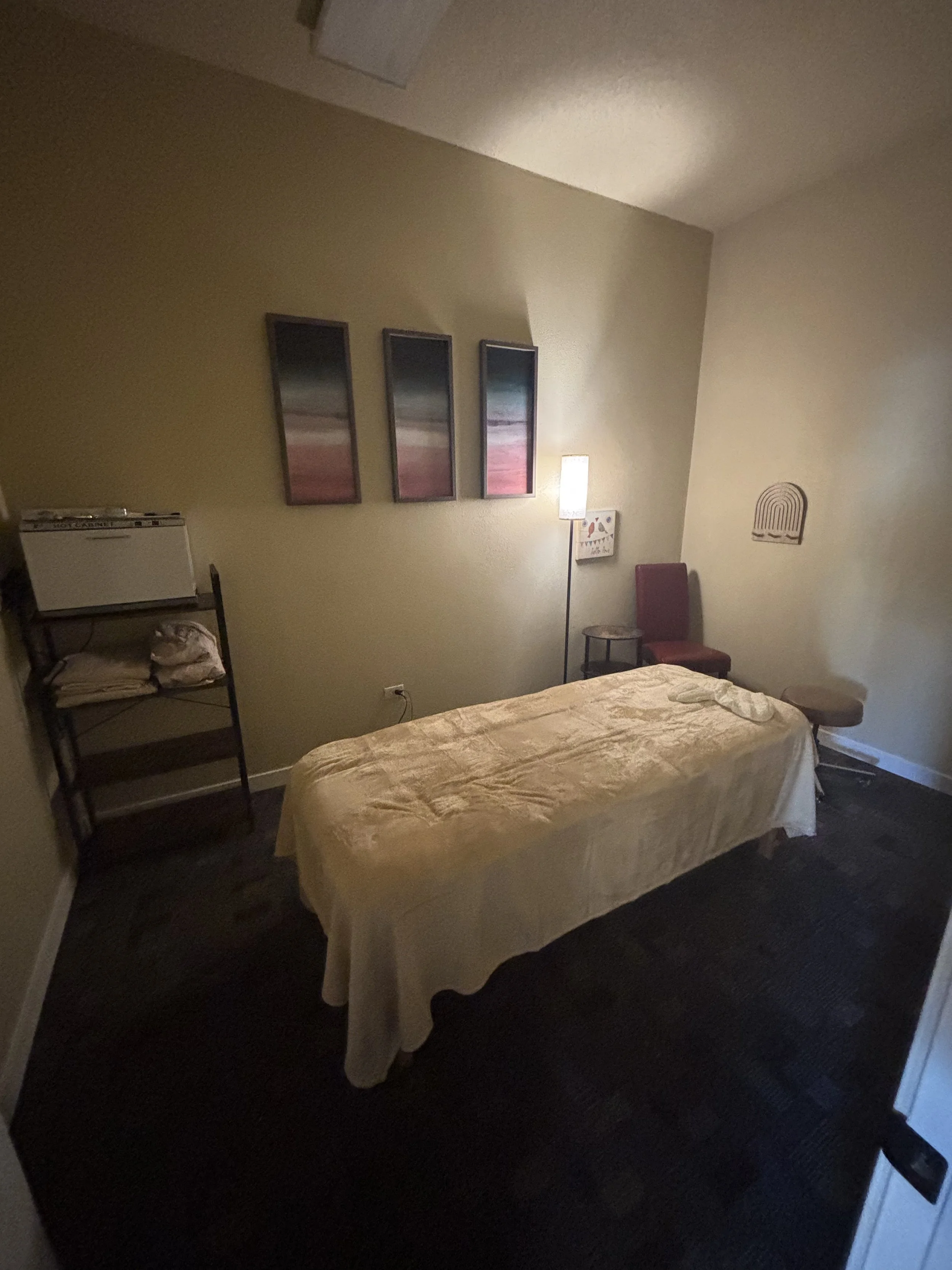 A massage room with a massage table covered in a beige sheet, a red chair, a small table, a floor lamp, a wall-mounted artwork, and a shelf with towels.