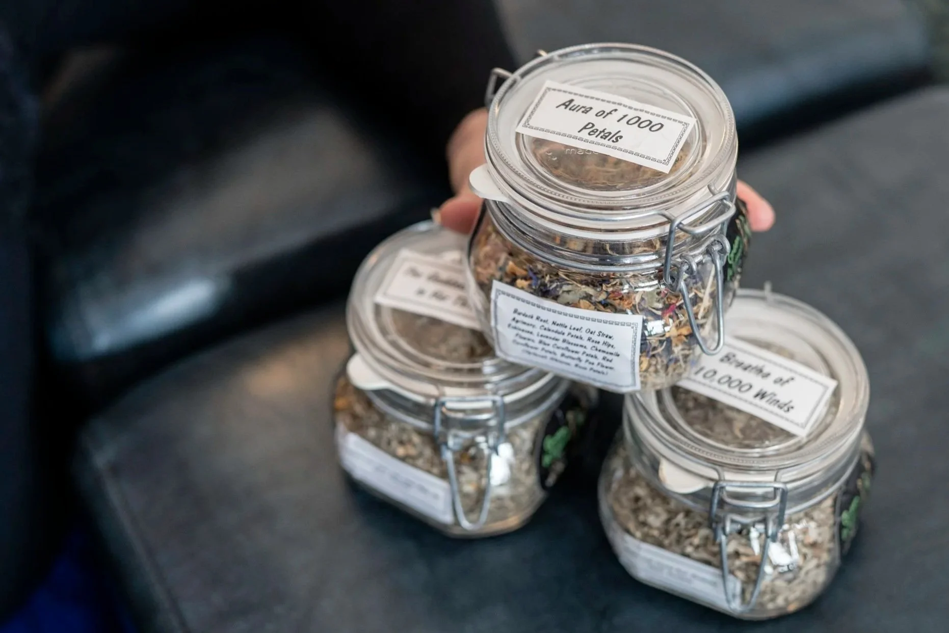 Three glass jars with labels and metal clasps on a dark surface, containing dried herbs and flowers.