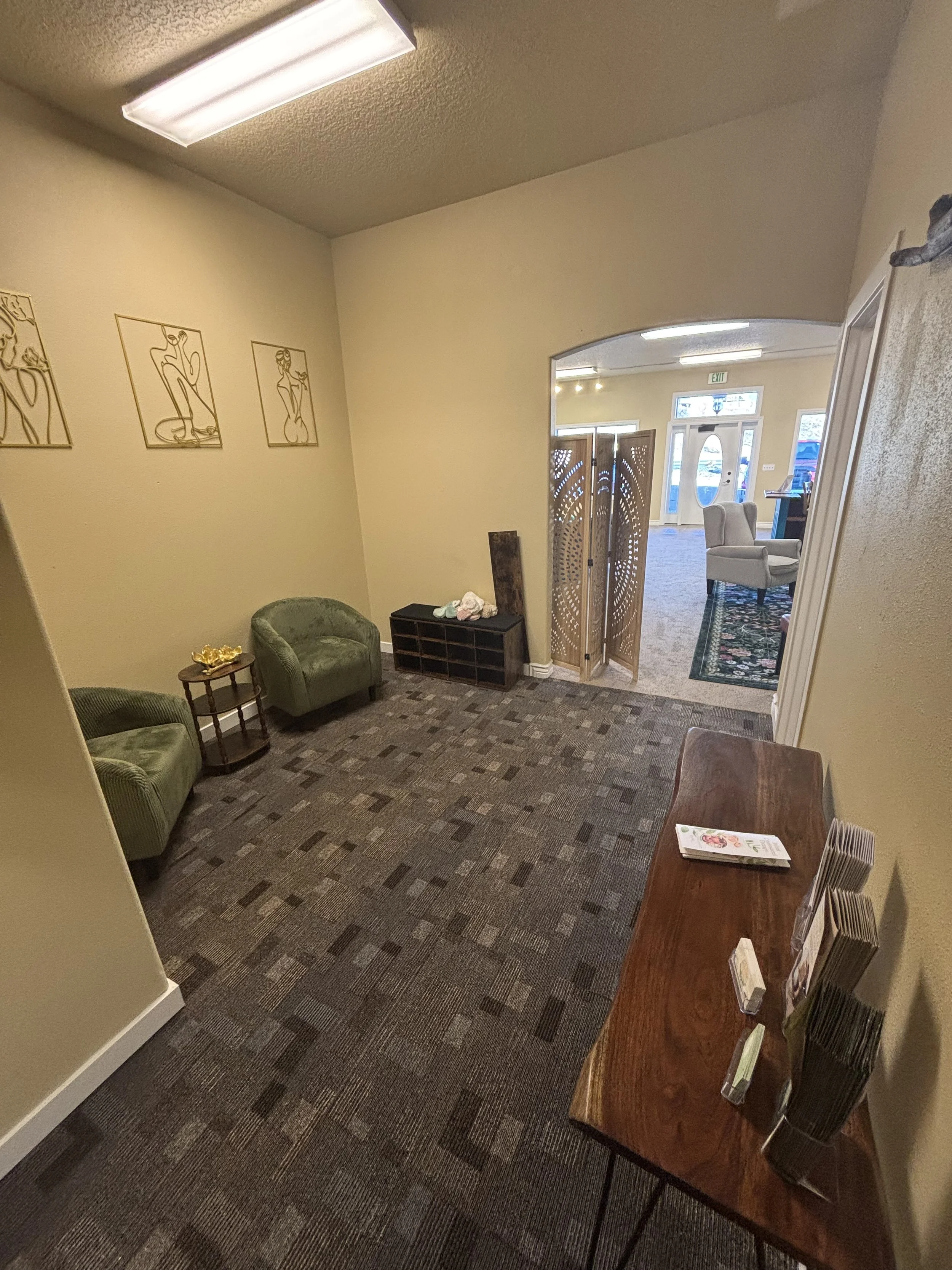 Small waiting area with two green chairs, a small wooden side table, a wooden magazine rack, a wooden console with brochures, and a decorative screen. The room has beige walls, a patterned carpet, and wall art of abstract figures. An open doorway lea