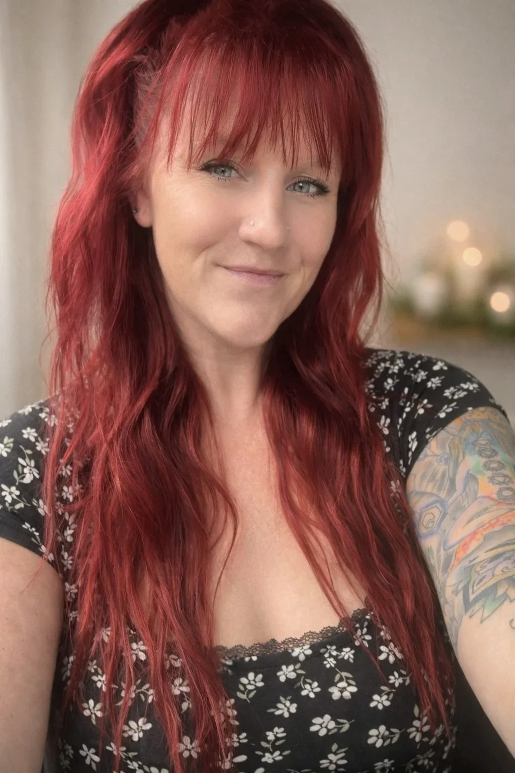 A woman with long red hair and a black floral dress, smiling at the camera, with a blurred background featuring some lights.
