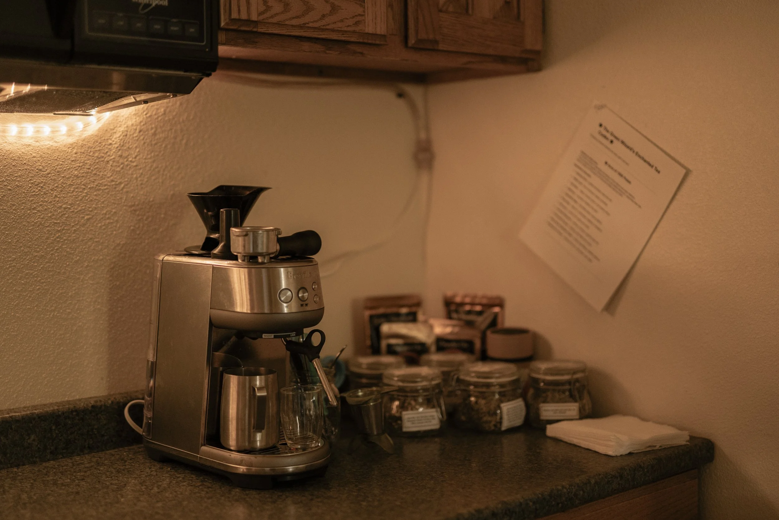 A coffee station with a stainless steel espresso machine, glass cups, jars of coffee beans, and a stack of napkins on a dark countertop in a cozy, dimly lit room.