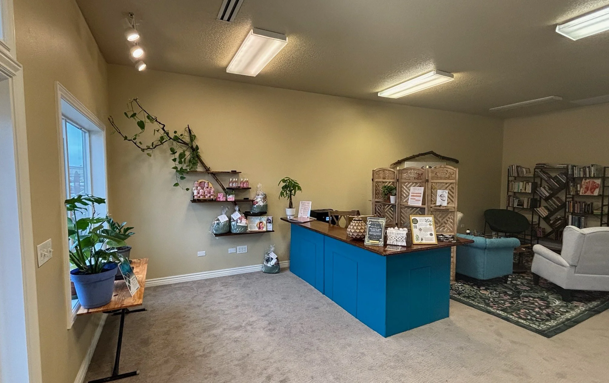 Interior of a cozy room with a reception desk, shelves with decorative items, plants, and a sitting area with armchairs and bookshelves.