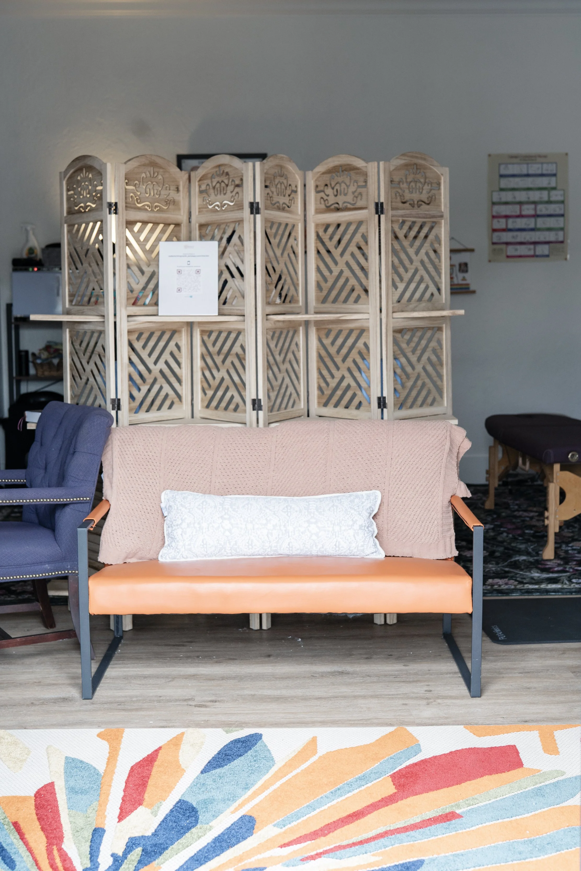 A room with a wooden privacy screen, a peach-colored bench with a white pillow, a blue armchair on the left, and a colorful rug on the floor.