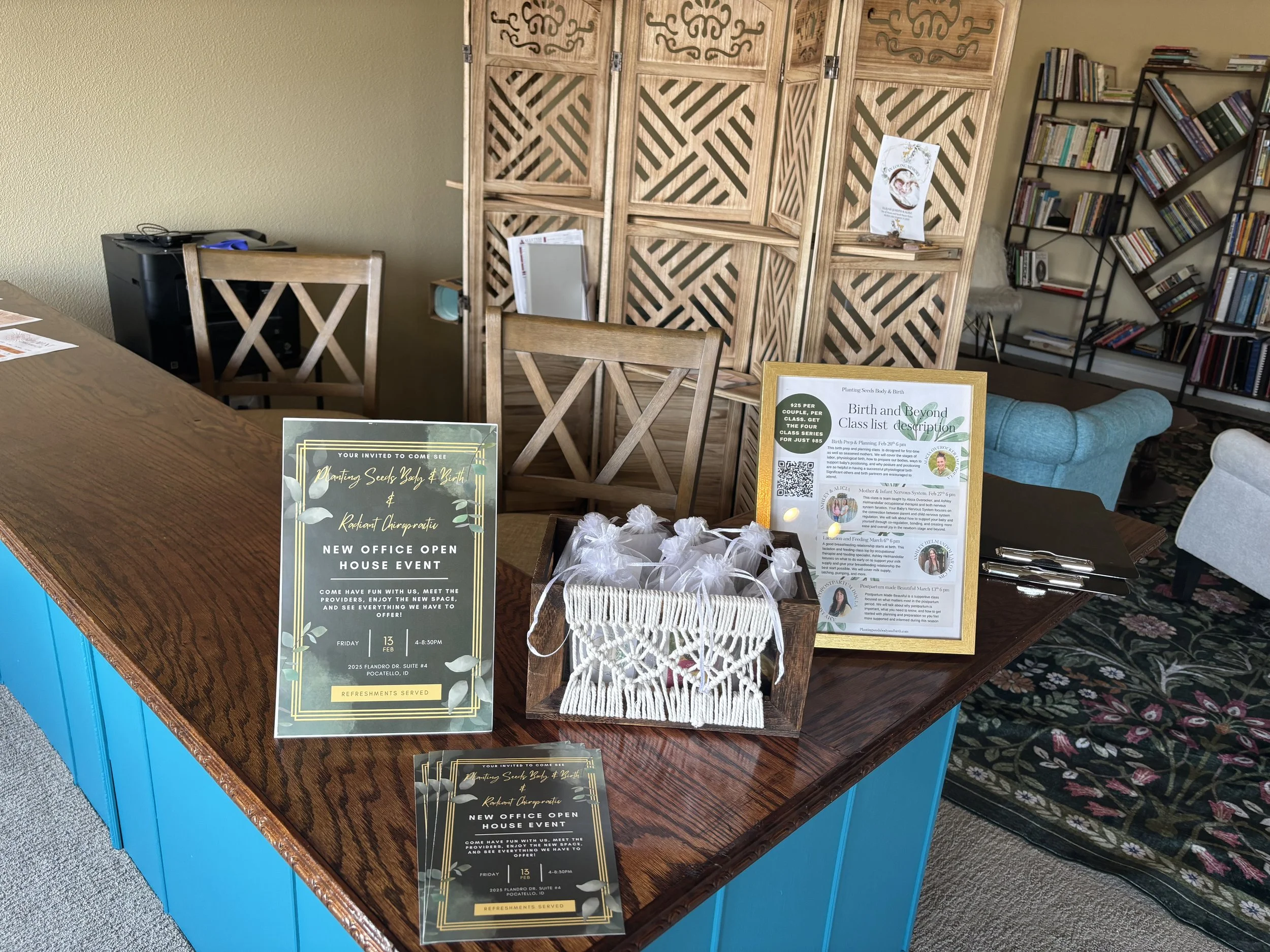 A wooden table with informational flyers, small gift bags in a basket, and pens, set in a cozy room with bookshelves, chairs, and a decorative folding screen.