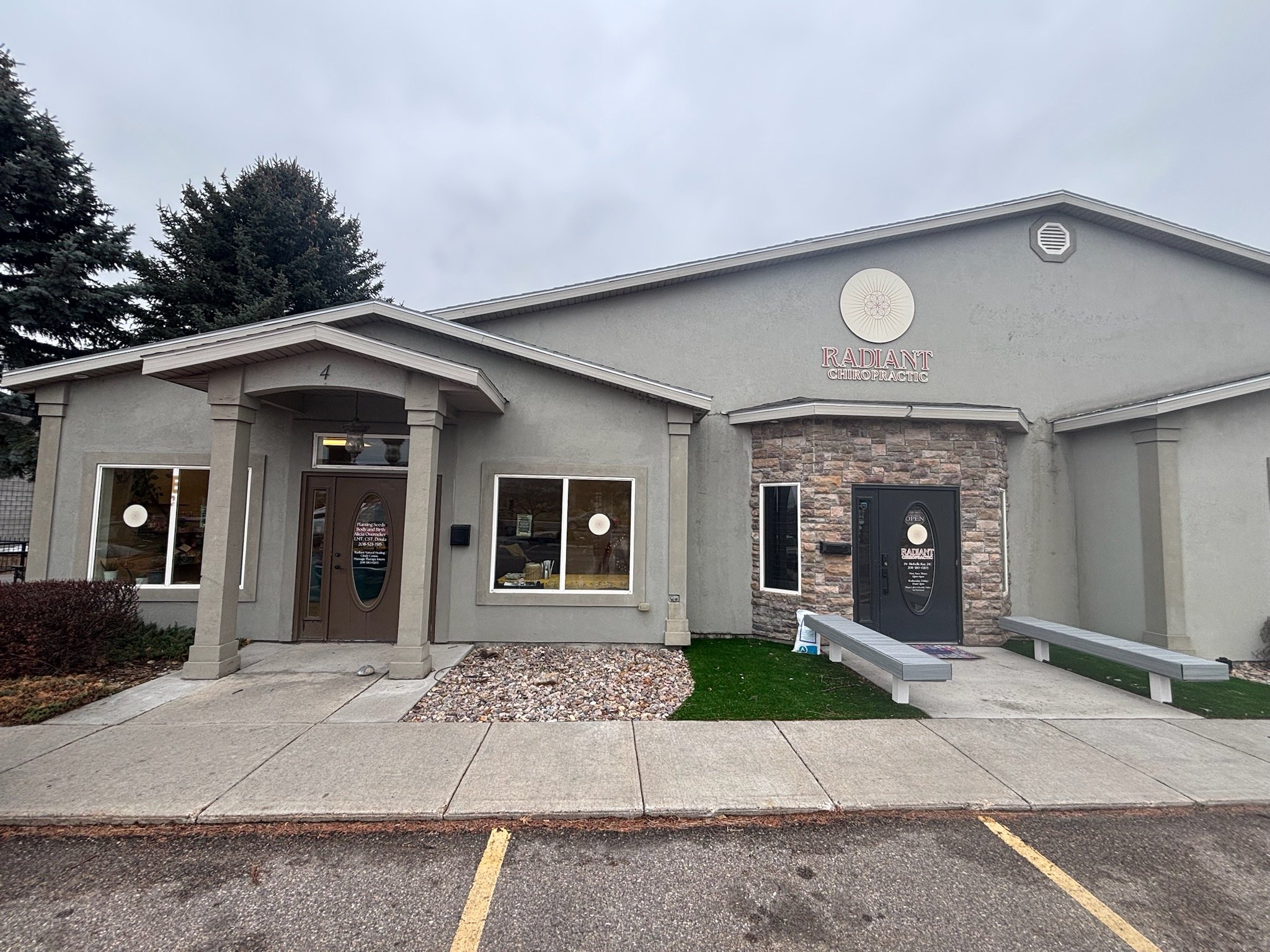 Exterior view of a chiropractic office called Radiant Chiropractic with a gray facade, two entrances, and parking spaces in front. The building has a stone accent wall near the entrances and signs displaying the business name.