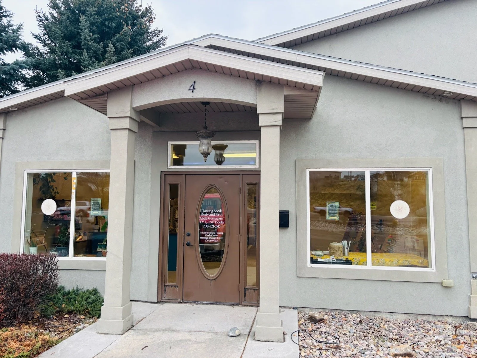 Front entrance of a commercial building with the number 4 above the door, featuring a brown oval-paneled door, two large windows on each side displaying items inside, and an outdoor light fixture hanging under the porch roof.