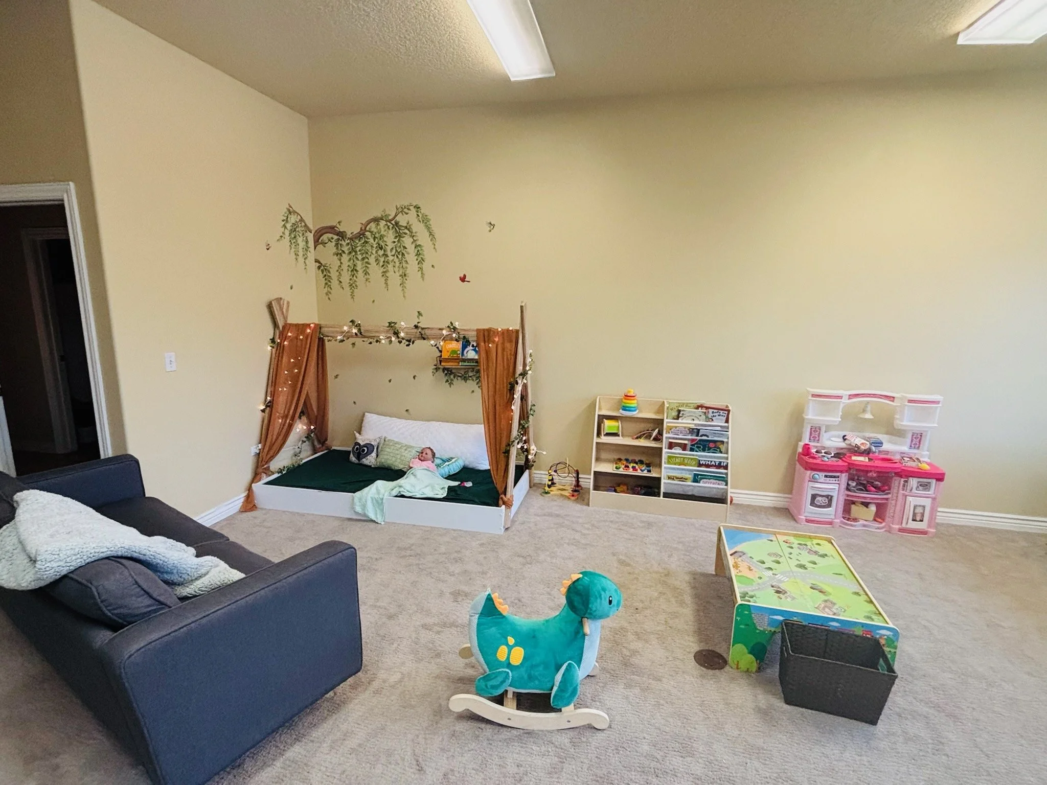 Children's playroom with a black sofa on the left, a wooden bed with drapes and fairy lights, a plush dinosaur rocker, a storage bin, a small bookshelf, a pink toy kitchen, and a colorful play table, all set on a beige carpet.
