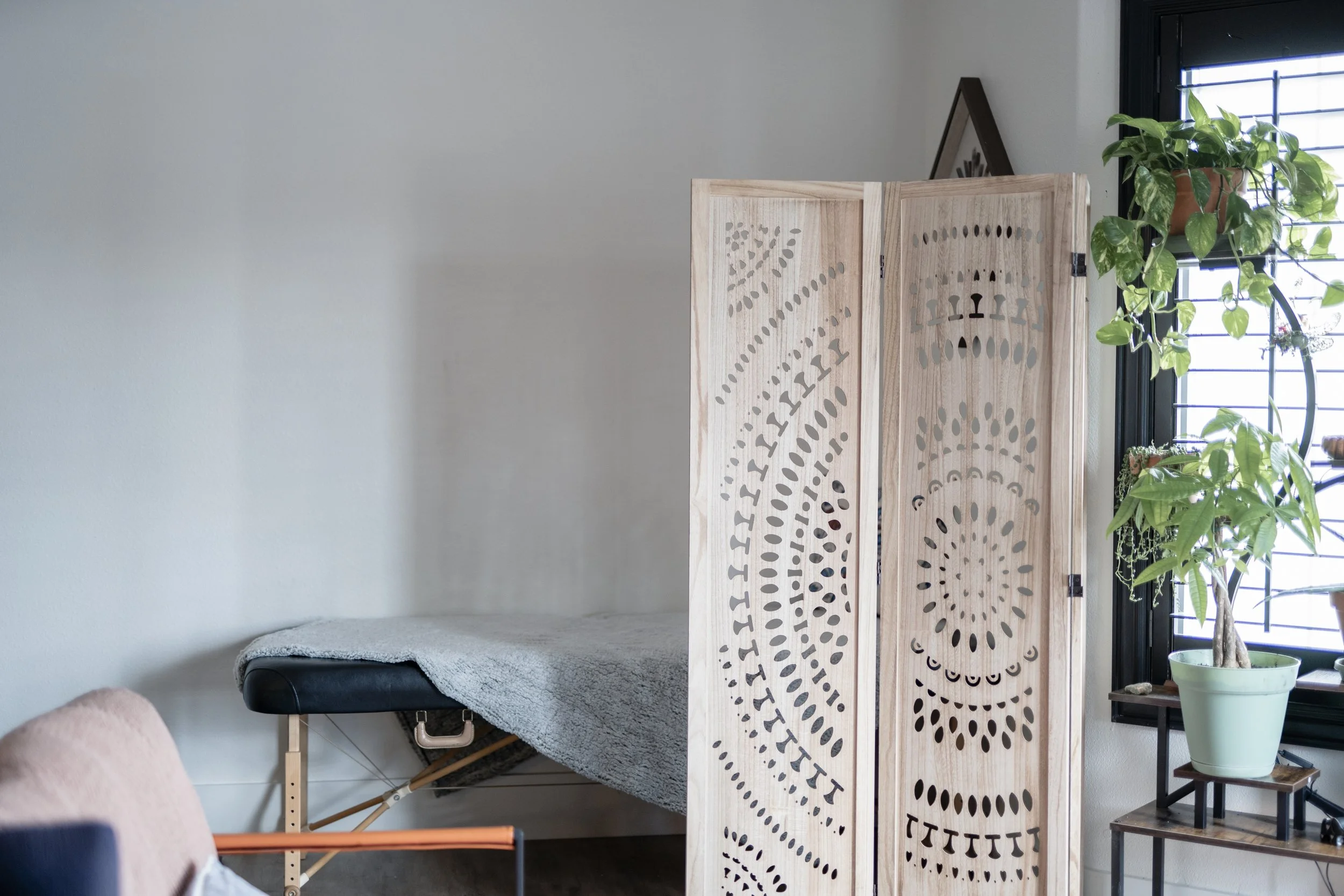 A room with a massage or treatment table covered with a gray blanket, a wooden decorative folding screen with cutout patterns, and a window with green plants in pots on a stand.