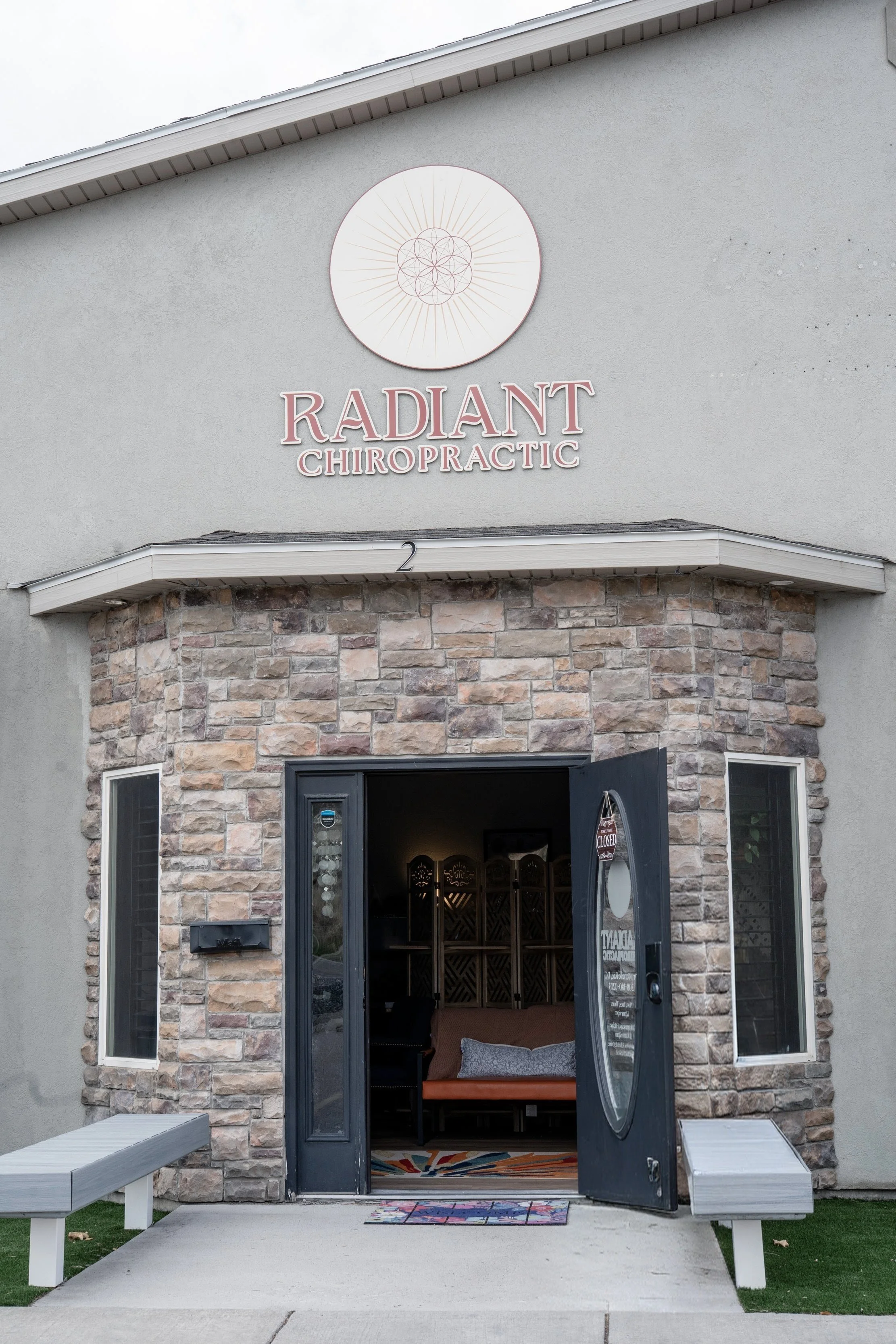 Exterior view of a building named Radiant Chiropractic with a stone facade and an open door, revealing a waiting area with a bench and colorful rug inside.