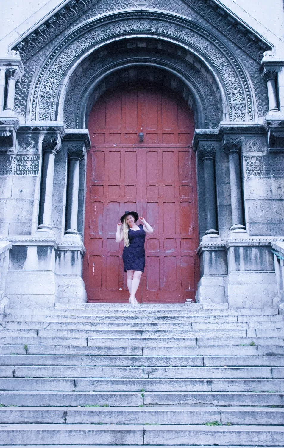 Woman in black dress and hat standing on stone stairs in front of large red wooden door with ornate stone archway, historic building.