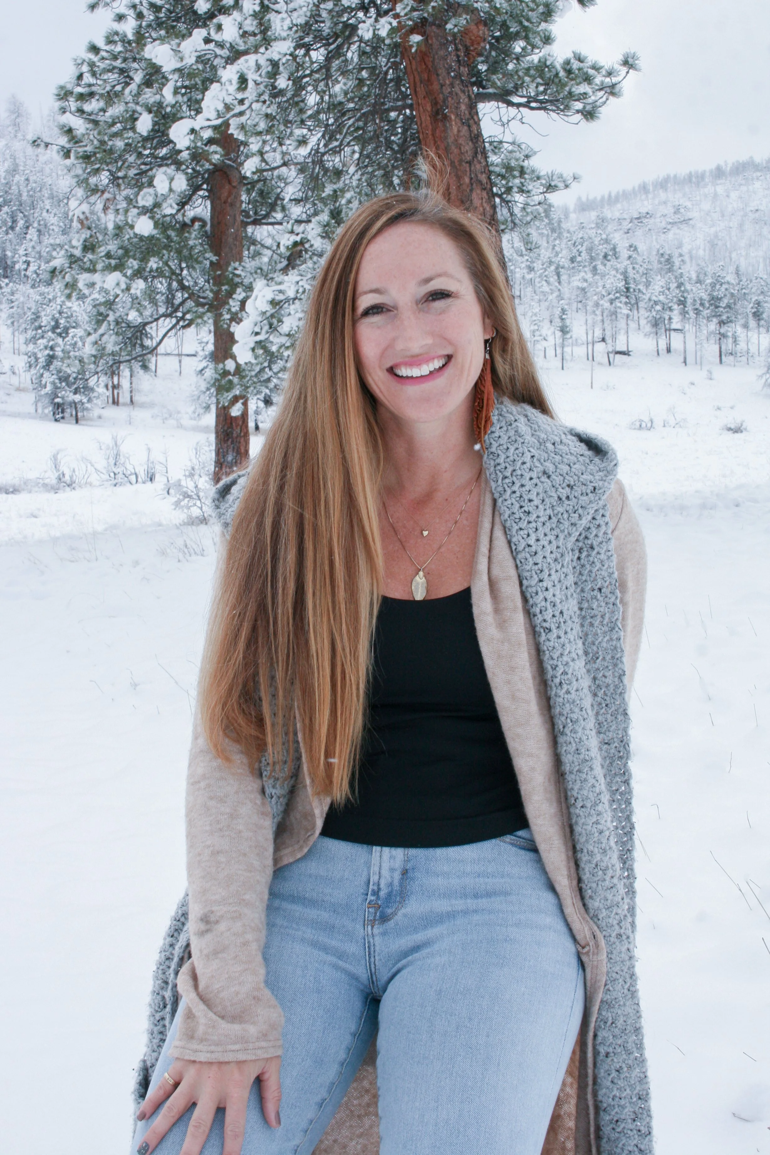 A woman with long red hair smiling outdoors in a snowy landscape with tall pine trees.