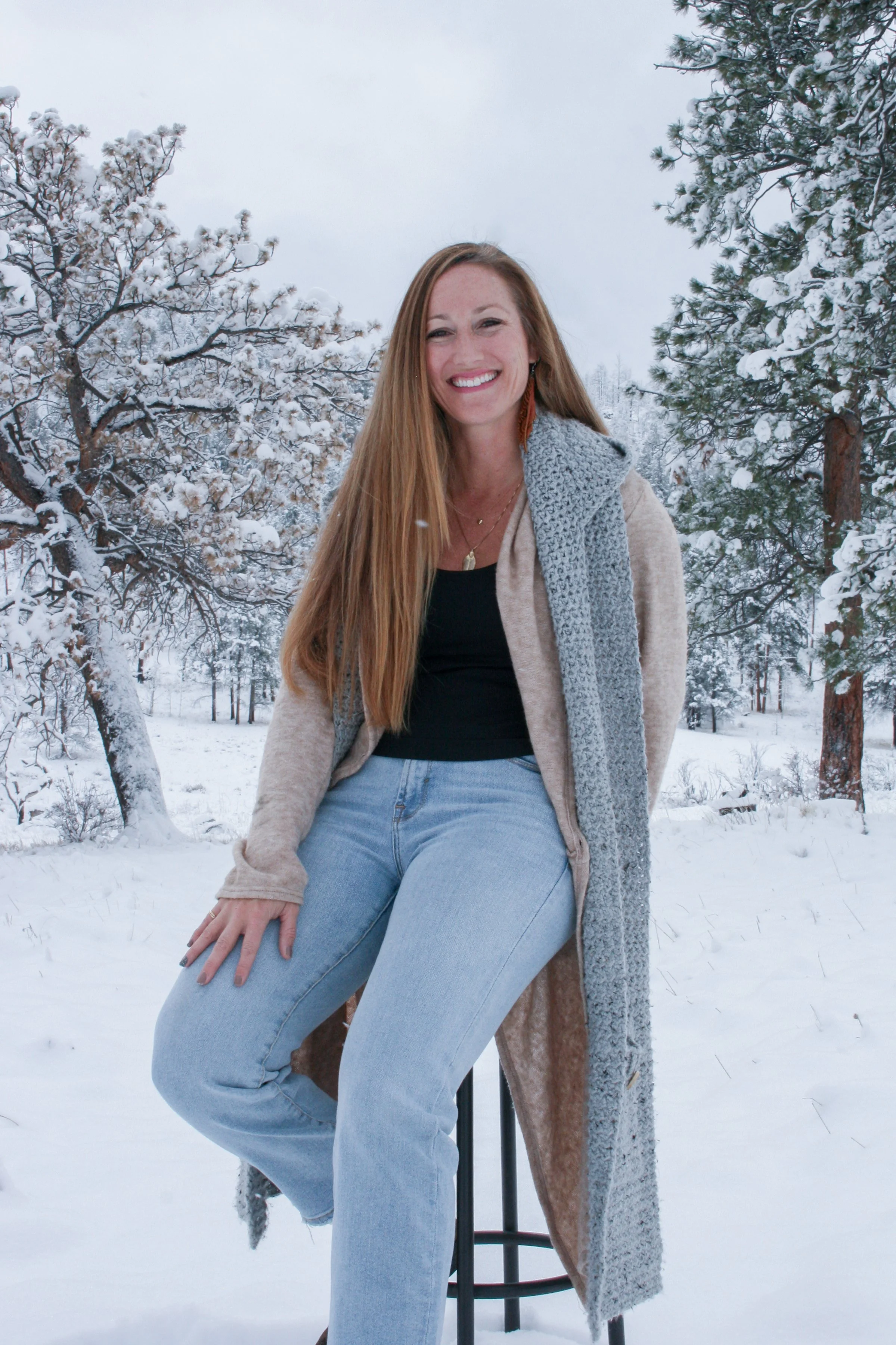A smiling woman with long red hair sitting outdoors in a snowy landscape with snow-covered trees, wearing a long beige coat, a black top, and light blue jeans.
