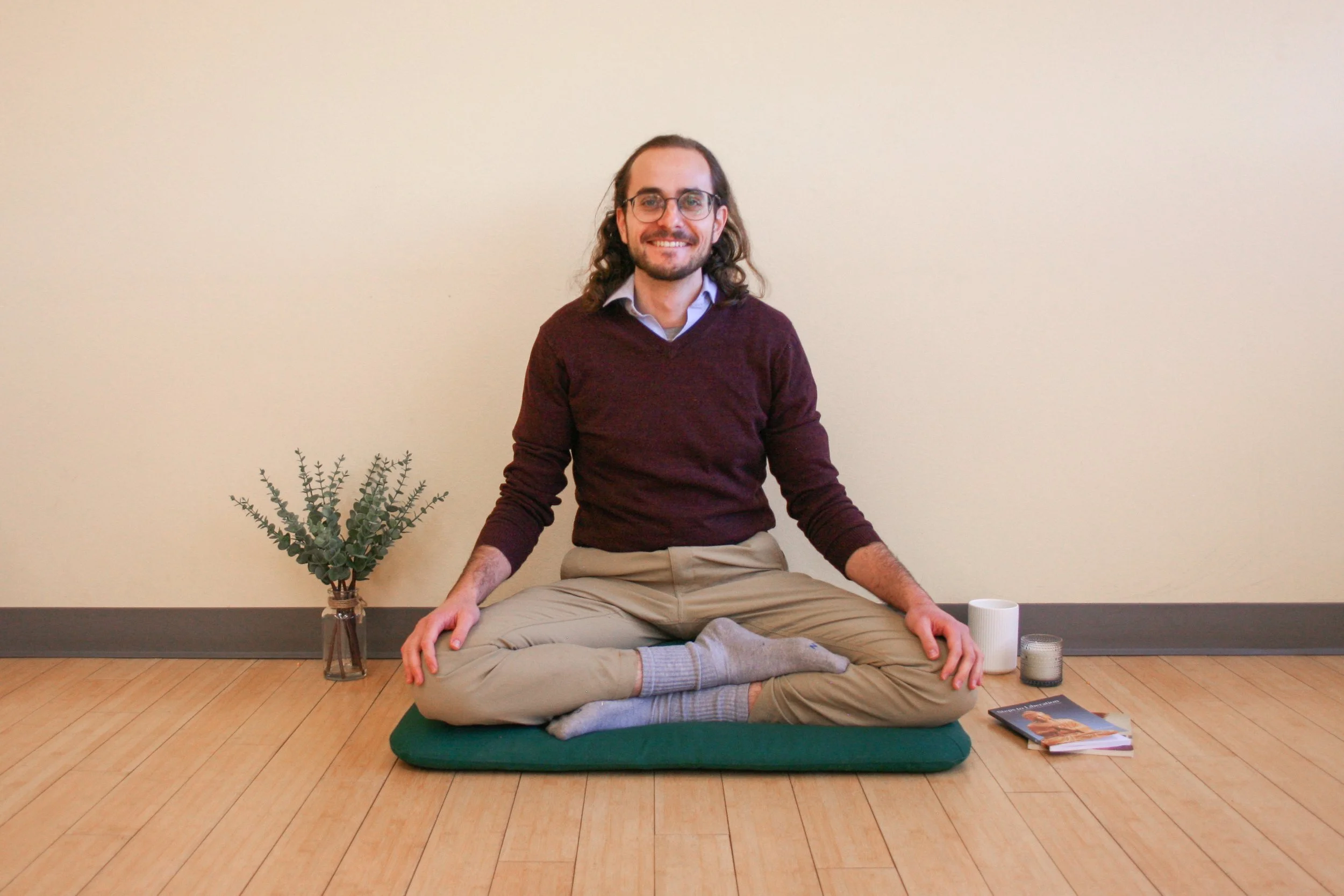 A man with long hair and glasses smiling in a seated yoga pose on a green cushion on a wooden floor, against a plain beige wall, with a small plant in a glass jar, a white mug, a gray container, a gray candle, and magazines nearby.