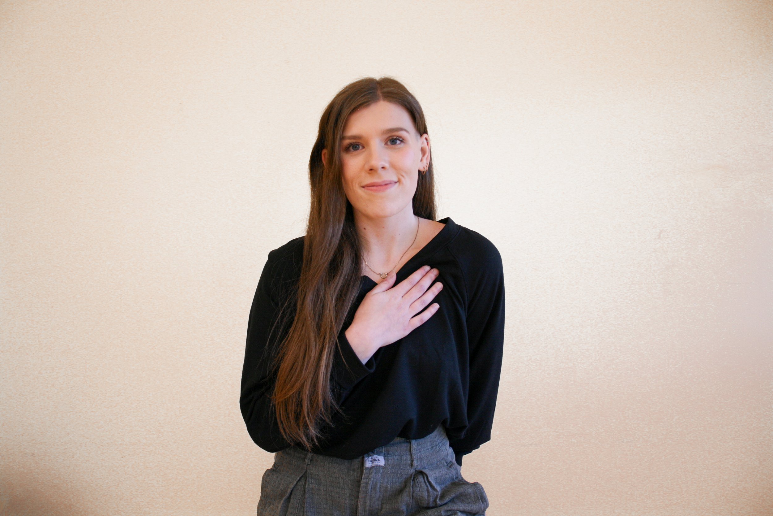 Young woman with long brown hair, wearing a black long sleeve top and gray pants, standing against a plain light-colored background, holding her hand over her chest with a gentle smile.