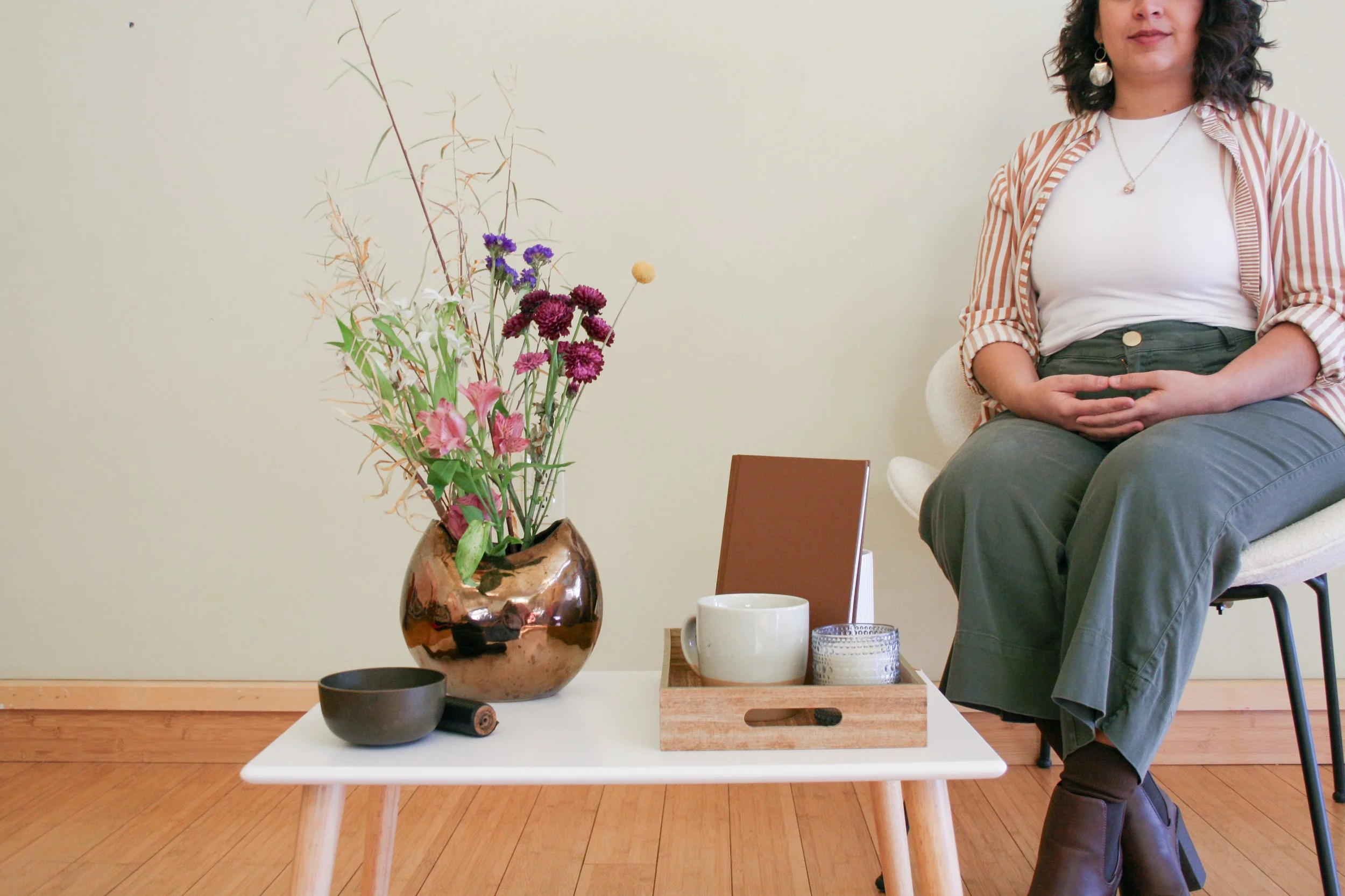A woman sitting on a white chair with a black metal frame, hands resting on her lap, wearing a striped shirt and dark green pants. In front of her, there's a small white table with a brown vase of colorful flowers, a black bowl, a white mug, and a small candle holder. The room has light green walls and wooden flooring.