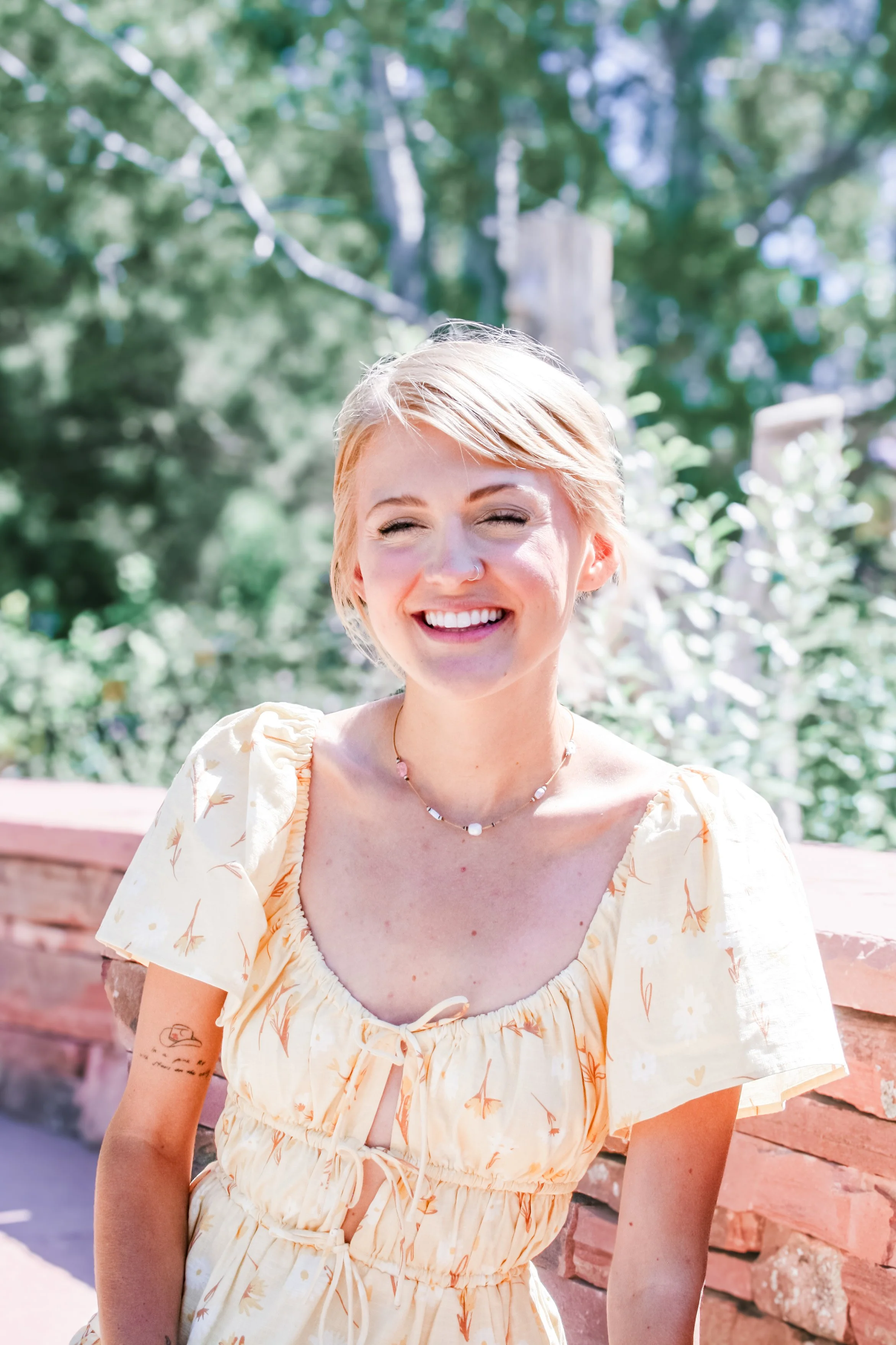 A smiling young woman with short blonde hair, wearing a light yellow dress with floral patterns, outdoors near a pink brick wall with trees in the background.