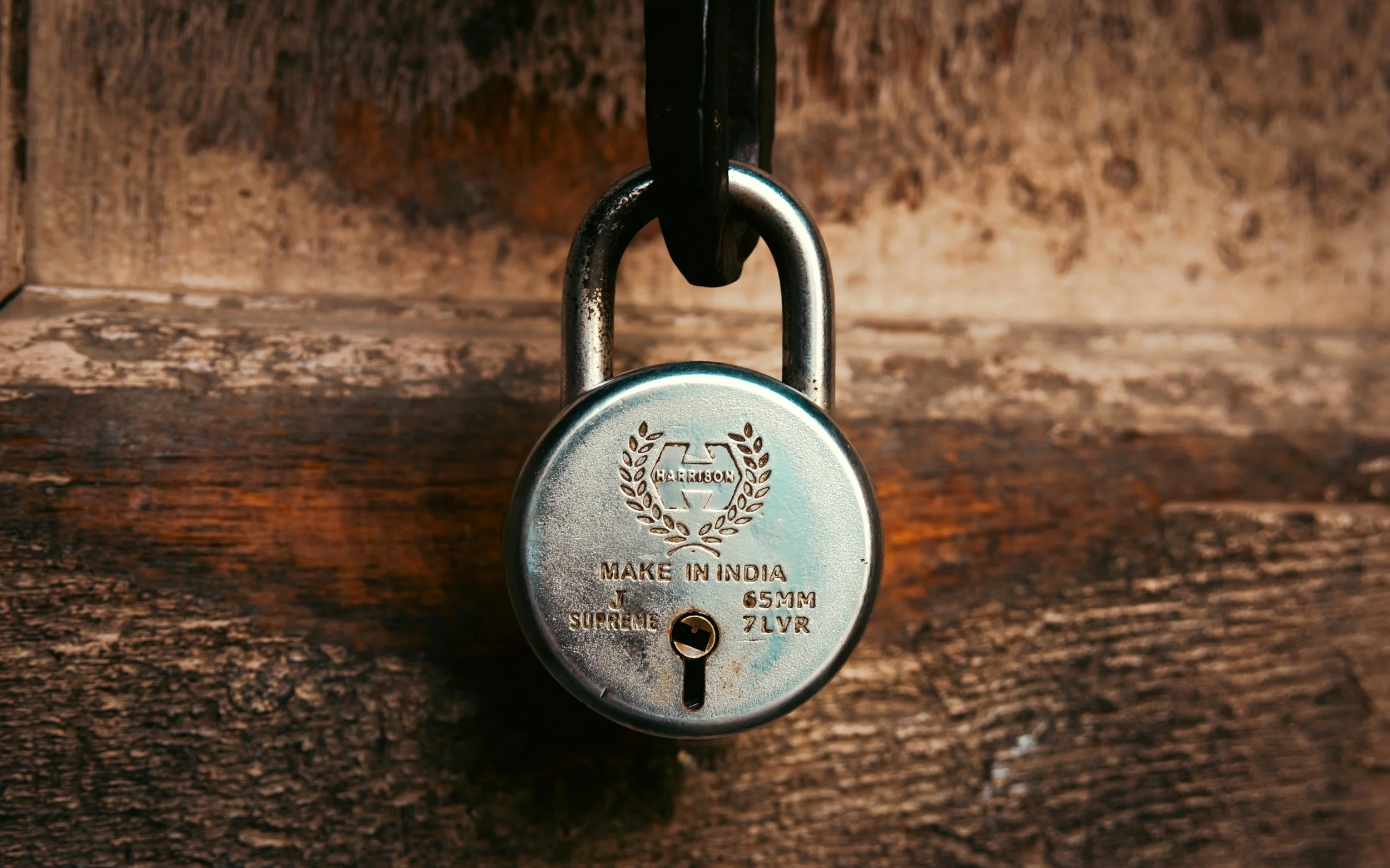 A silver padlock hanging from a black hook, attached to a weathered wooden surface, with engravings including 'HARRISON,' 'MAKE IN INDIA,' and other small text.