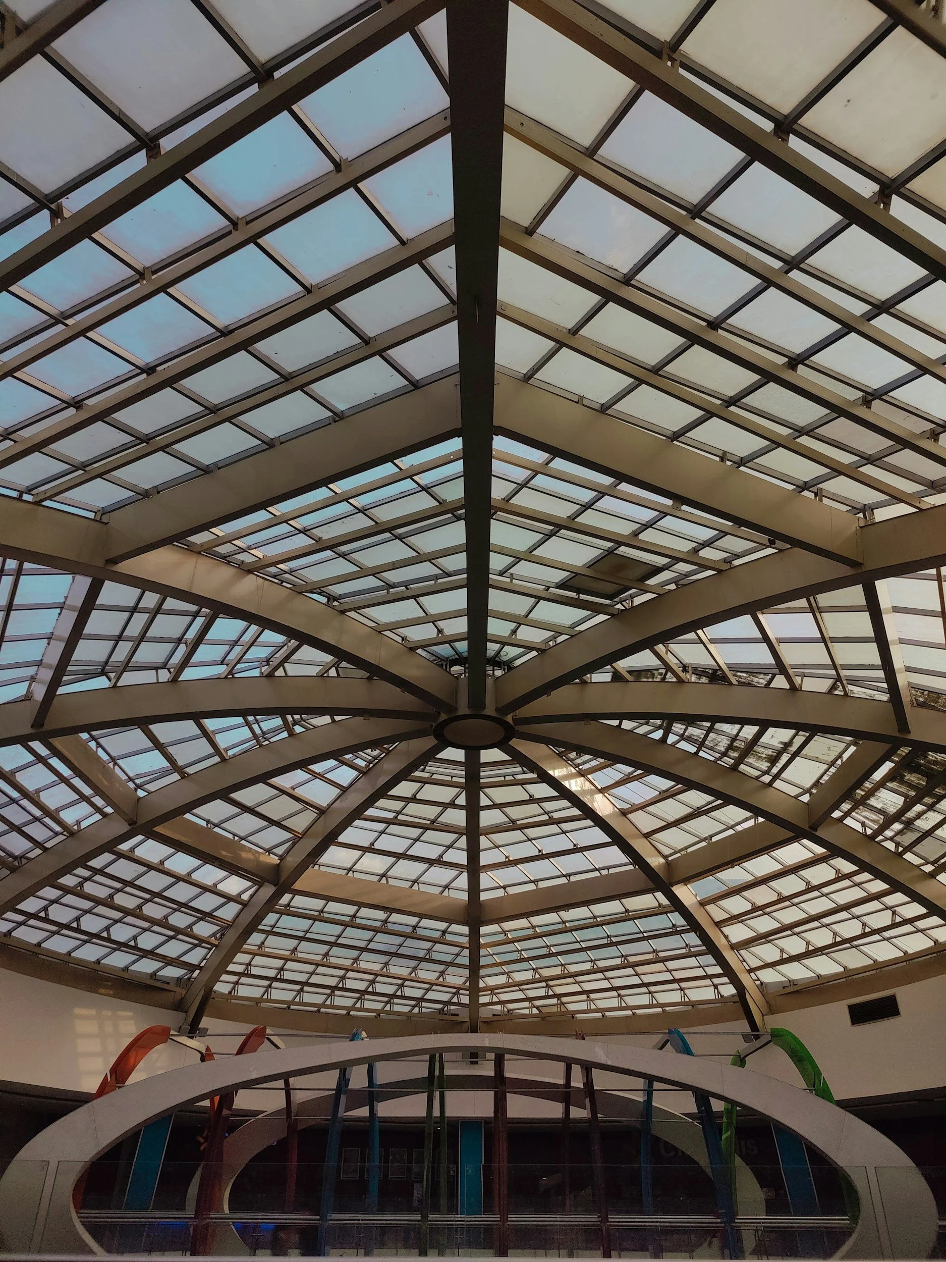Interior view of a large, glass-domed ceiling structure, likely in a shopping mall or public space, with supporting beams and a circular opening at the center.