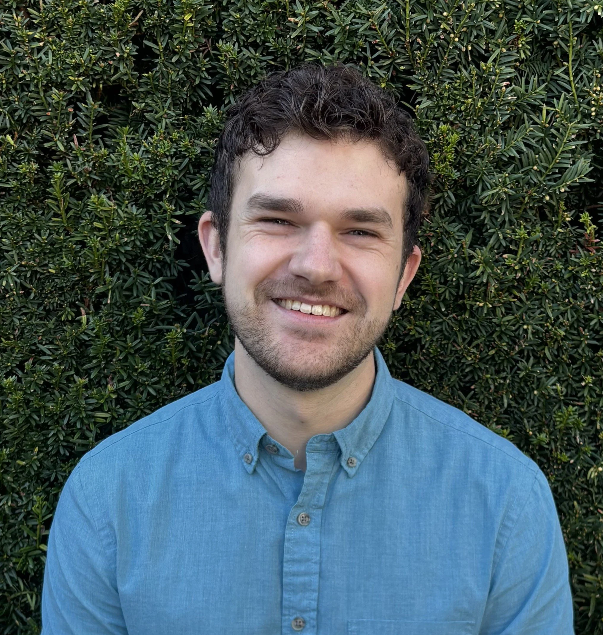 Photo of Brennan smiling in front of a green hedge