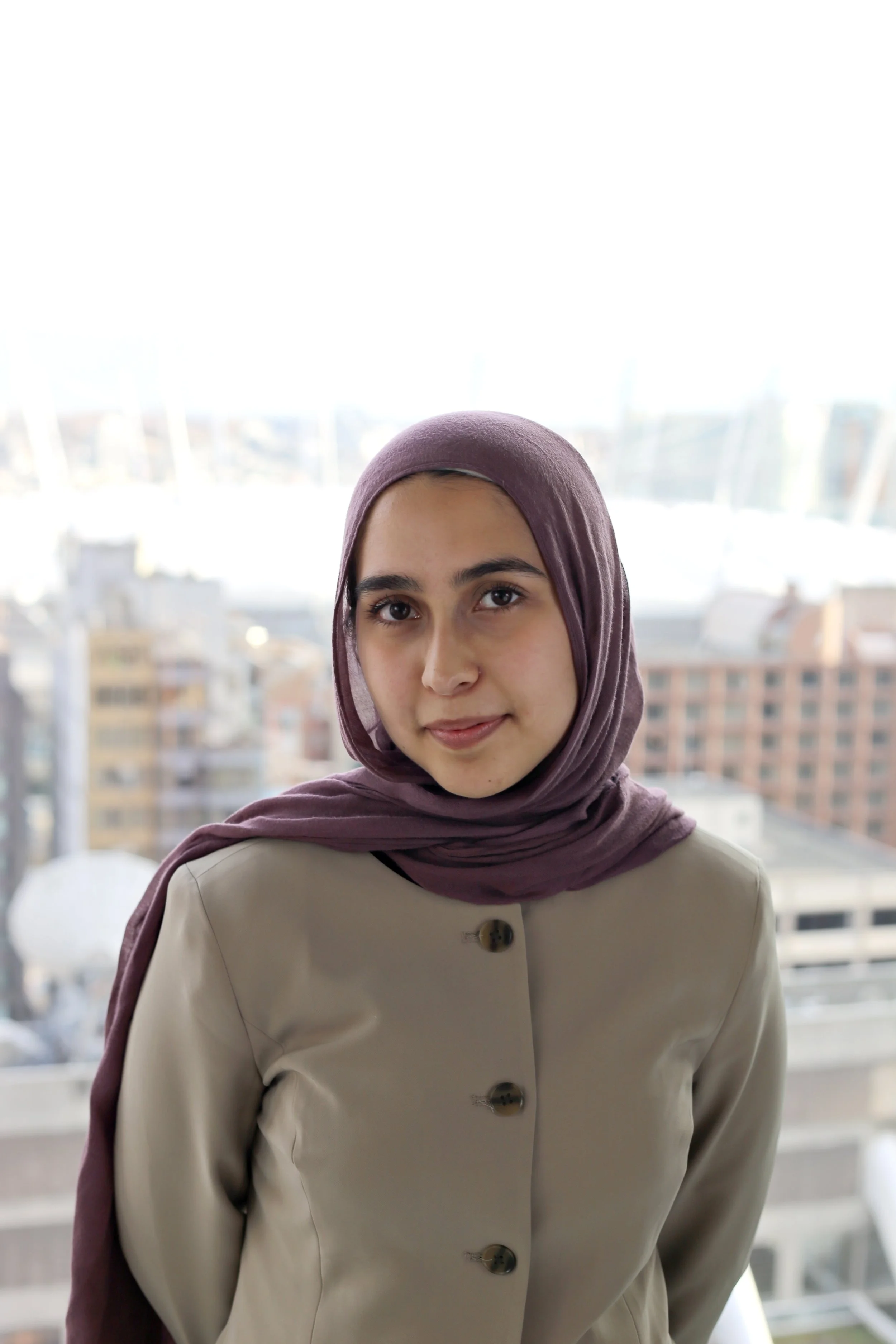 Farahnaz stands smiling in front of sunny skyline, wearing a a dusty pink headscarf and beige jacket.