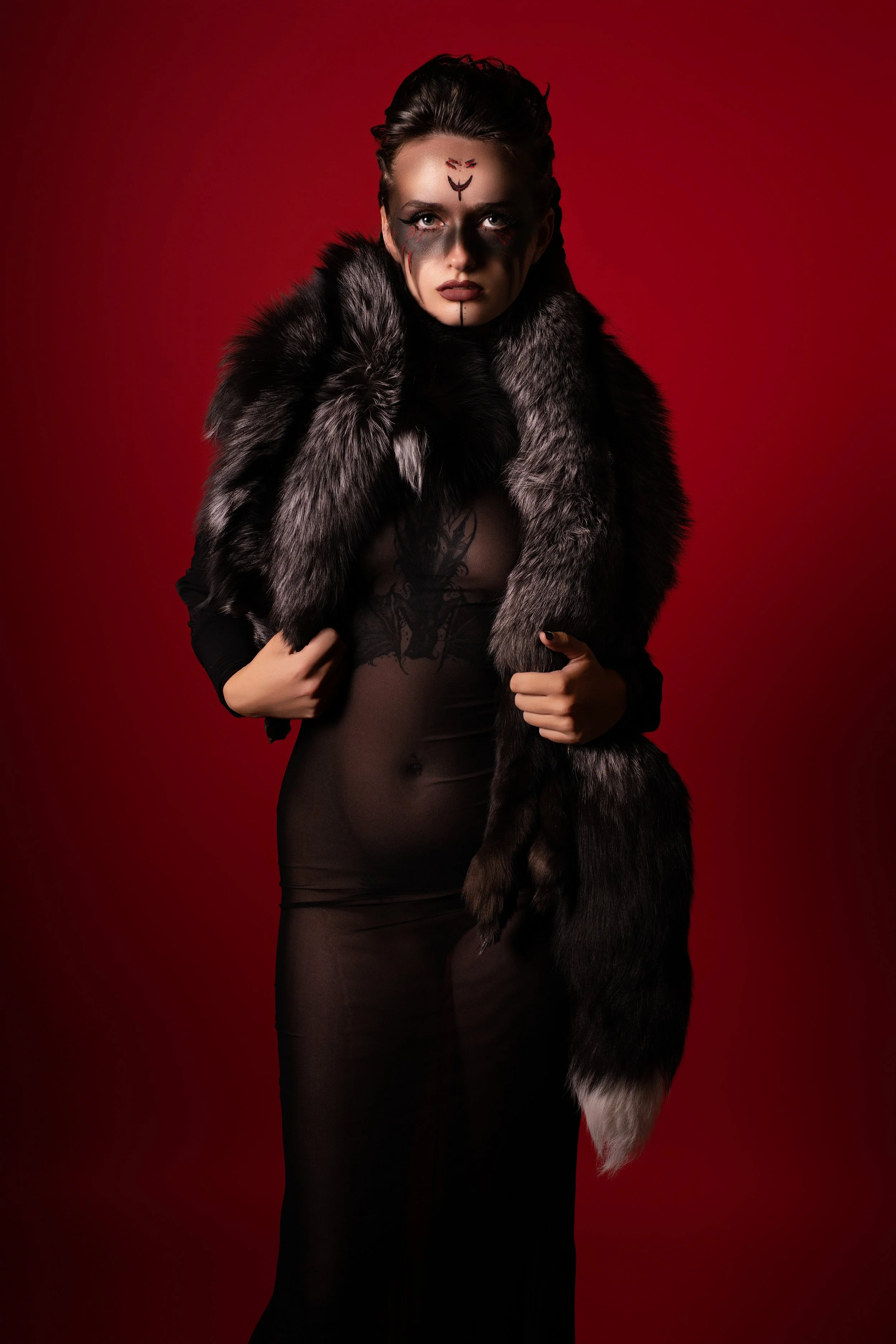 A woman with dark makeup and facial face paint, wearing a black sheer dress and a black and gray fur stole, stands against a red background.