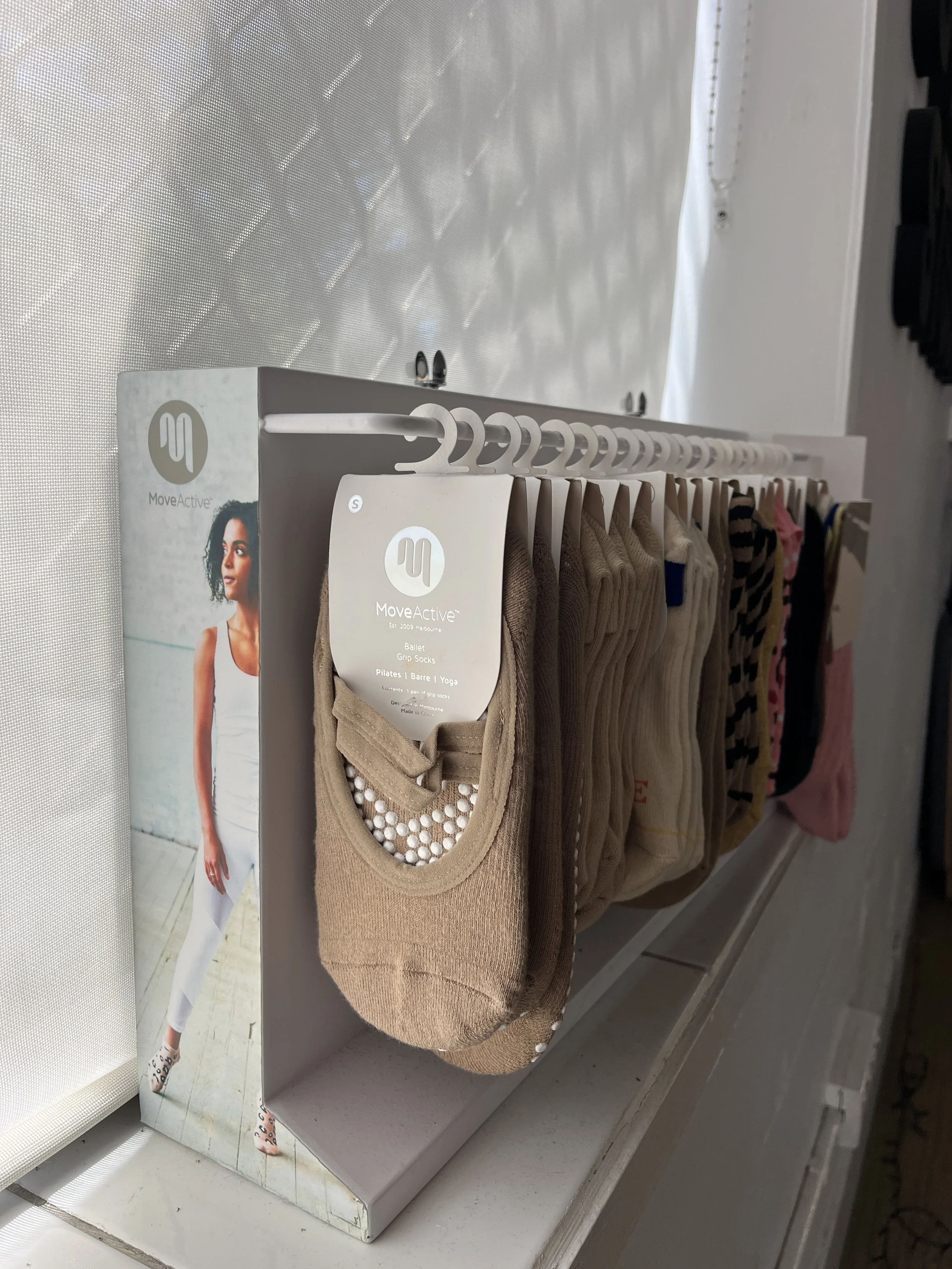 Display of various colorful socks hanging on a white rack with a MoveActive sign, with a promotional poster of a woman in white walking on a light-colored floor and wall in the background.