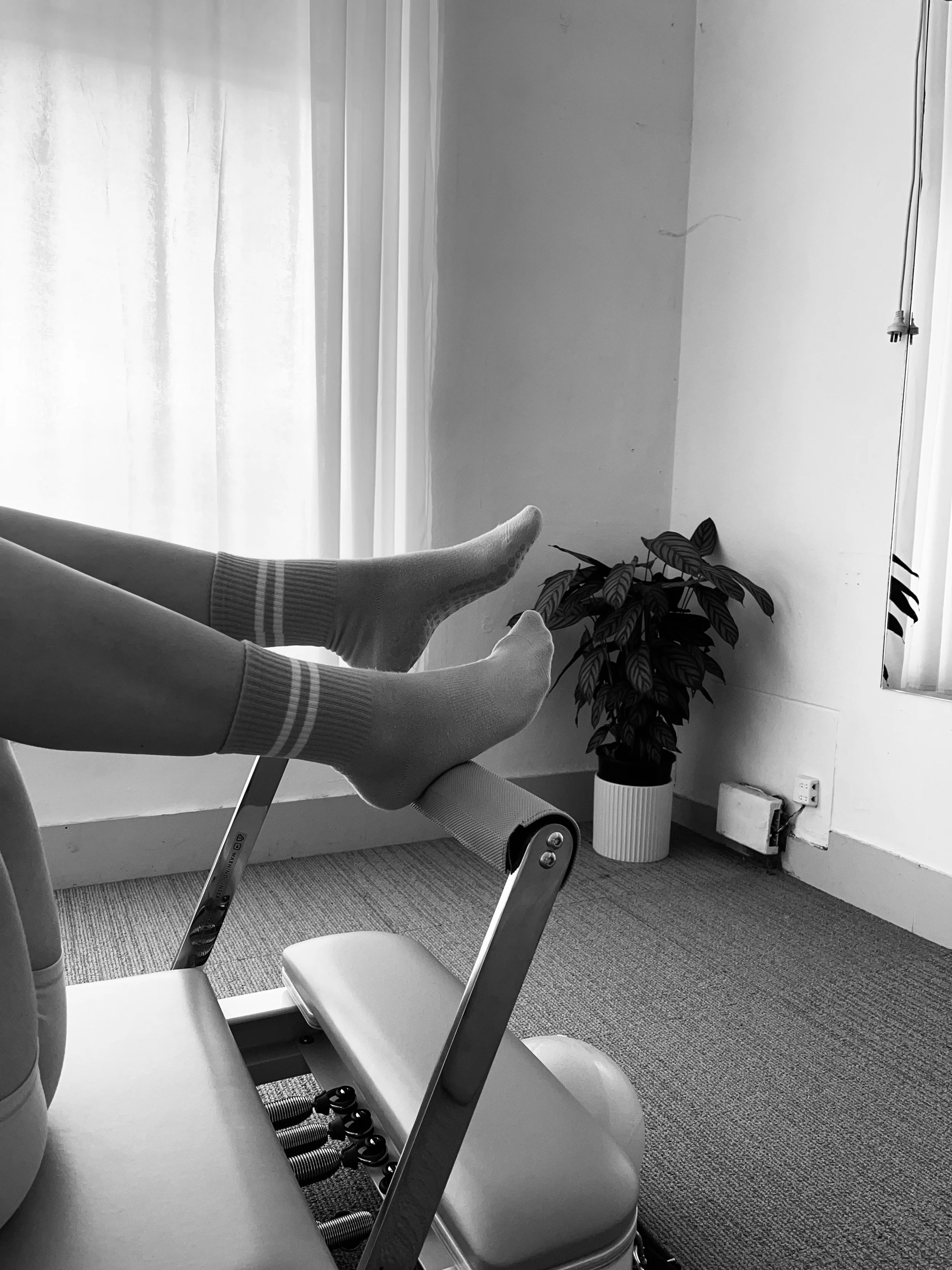 Person lying on a Reformer with legs stretched out, wearing socks. There is a potted plant near the wall, a mirror, and a window with curtains in the background.