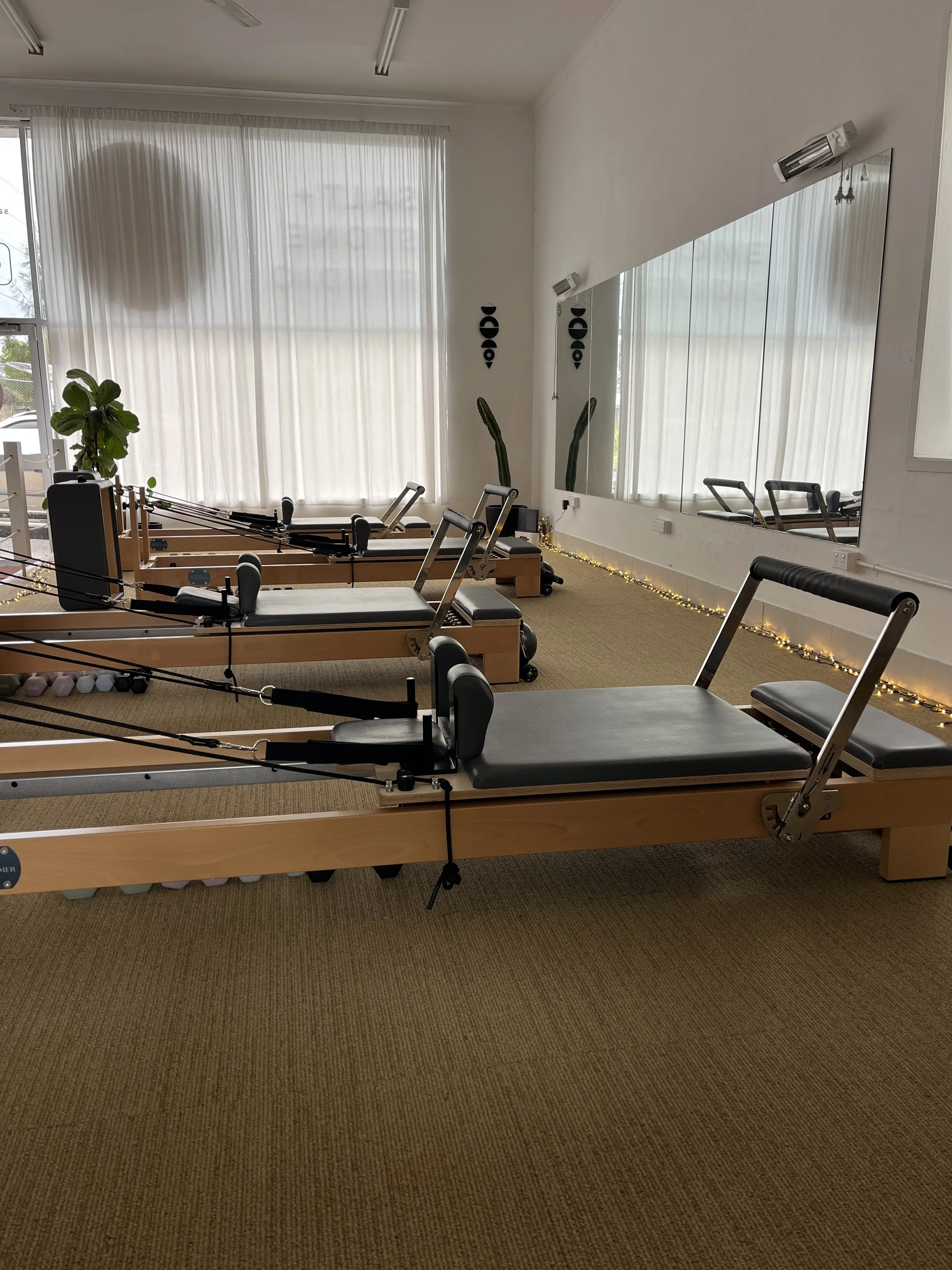 A Pilates studio with multiple reformer machines, large mirror, potted plants, and string lights along the wall, with white curtains covering large windows.